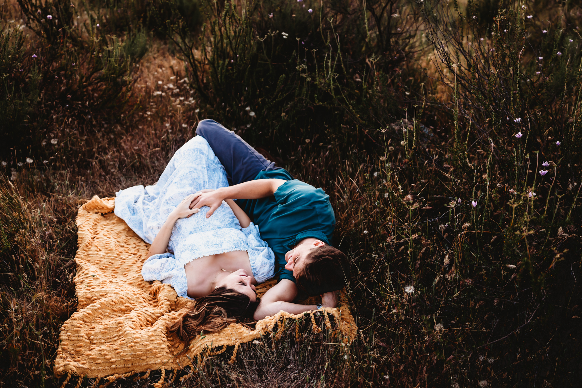 A pregnant woman wearing a long dress lays with her husband on a yellow blanket in a field of grass and wildflowers. This is during a San Diego lifestyle maternity photography session by Love Michelle Photography.