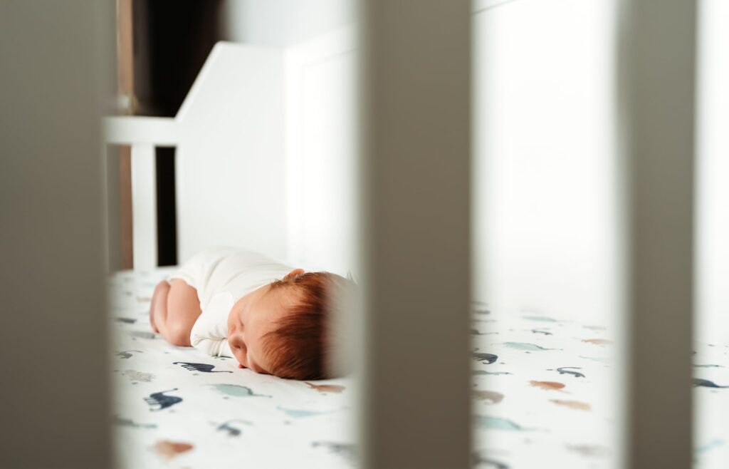 A red headed newborn baby boy wearing a white onesie sleeps in his crib during a lifestyle newborn photography session in San Diego. 