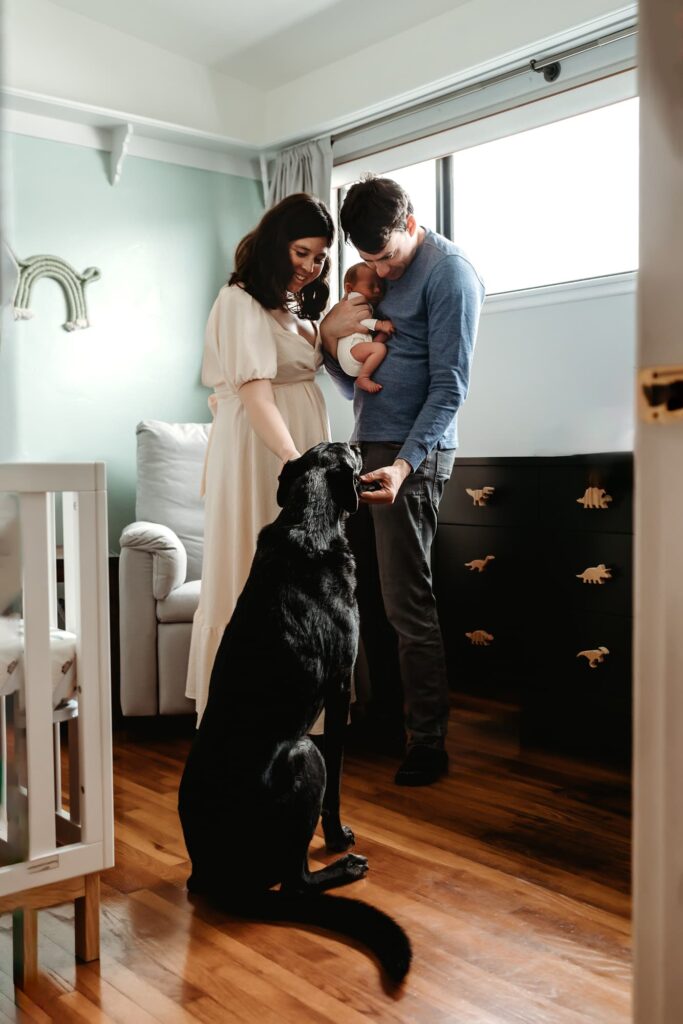 A couple holding their newborn baby boy look at and pet their black dog. 