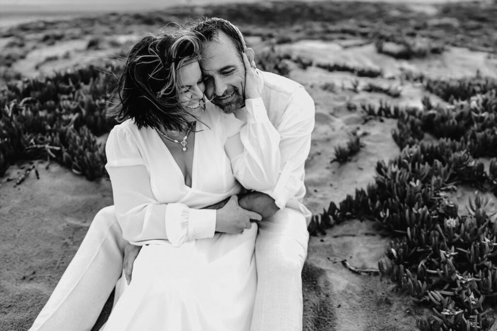 A husband and wife wear white and embrace each other while sitting on a Coronado Beach sand dune.