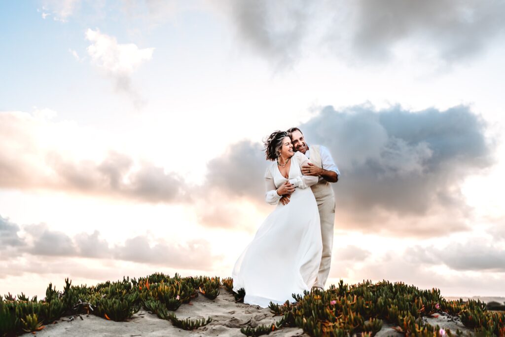 A man and his wife, both wearing white, stand on top of a San Diego beach sand dune, embracing and smiling.