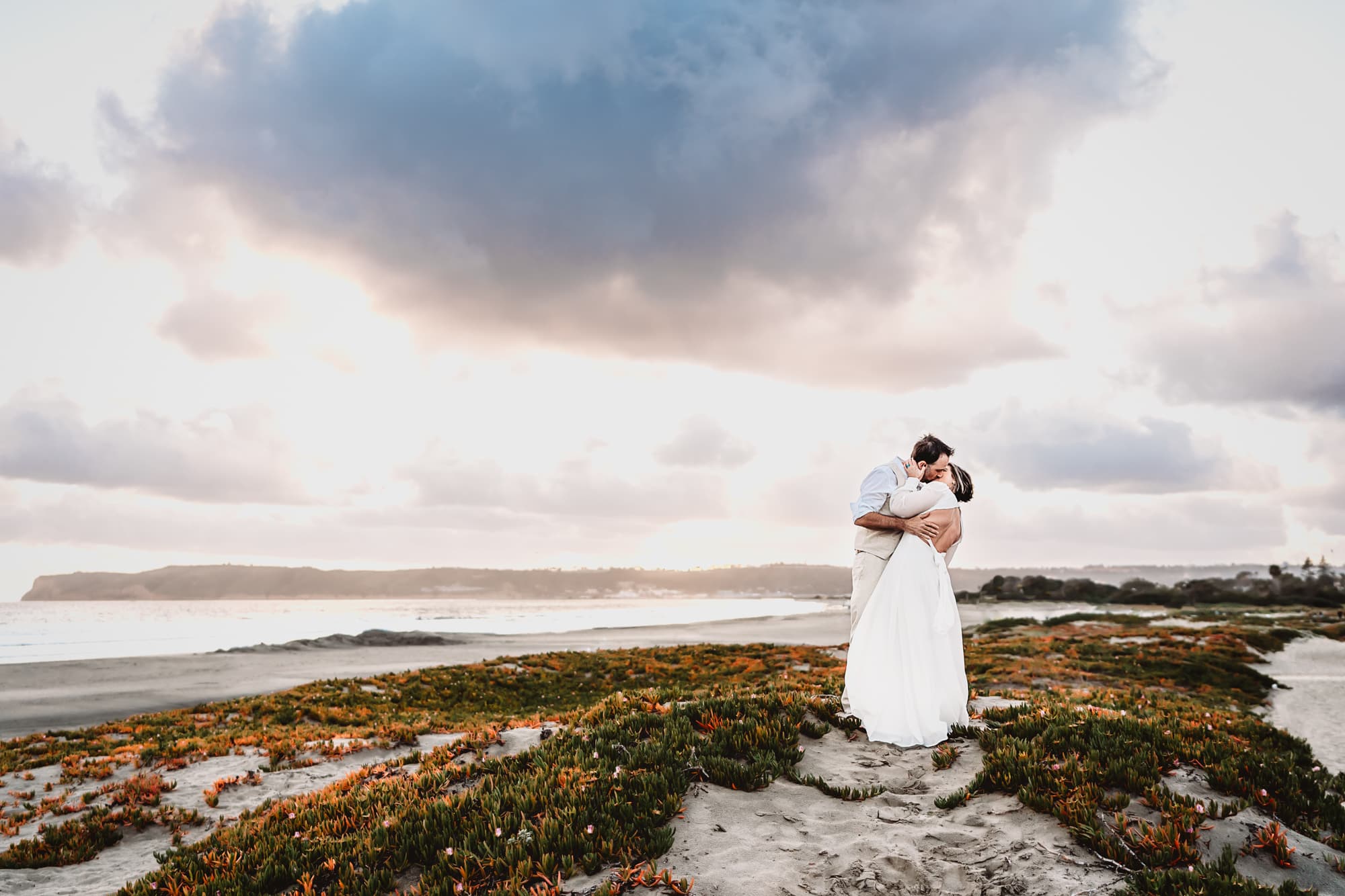 A man and wife stand and kiss one another during sunset on a Coronado Beach sand dune.
