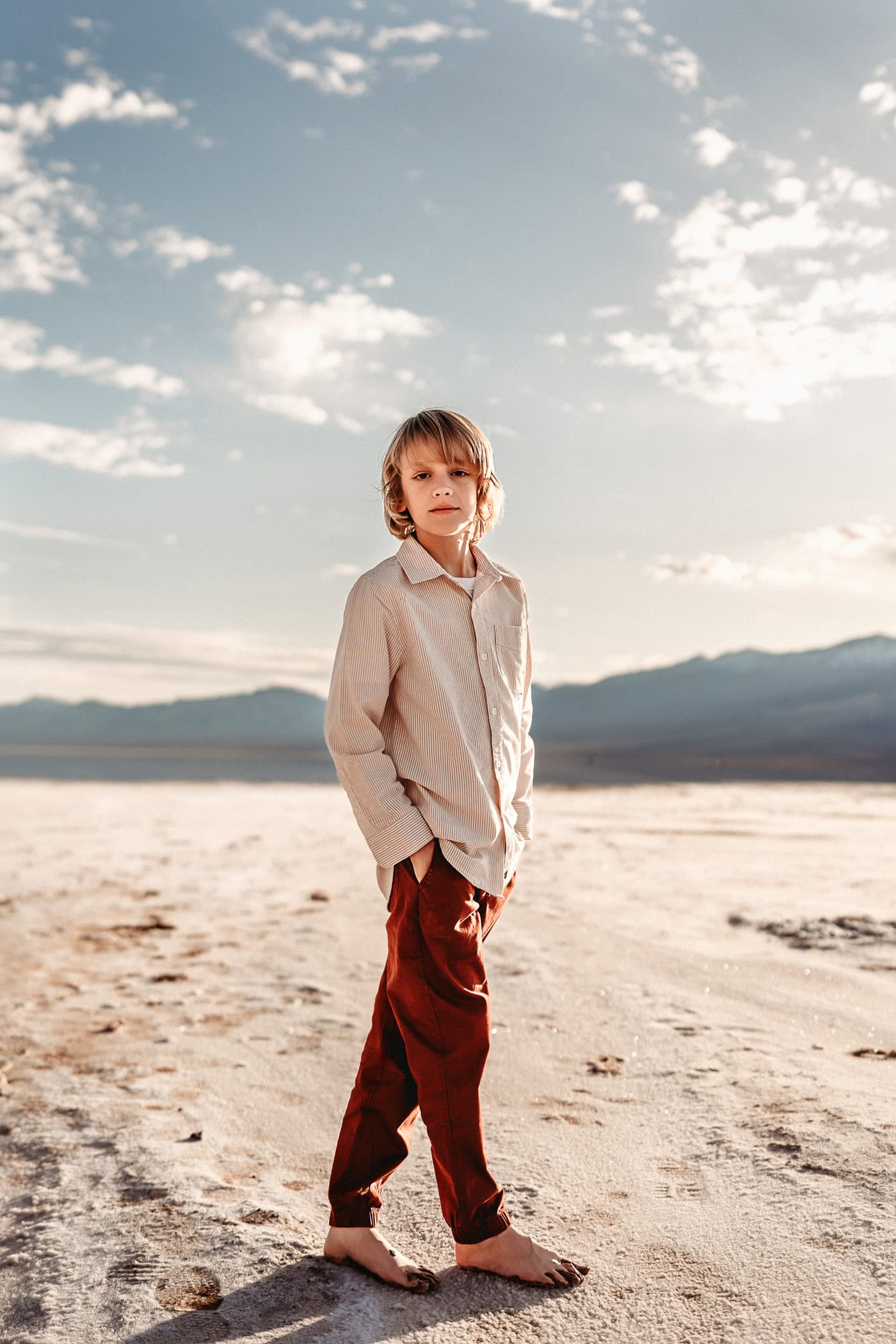 A ten year old boy stands in Death Valley National Park's Badwater Basin, barefoot, with is hands in his pockets, looking at the camera with a serious expression on his face. 