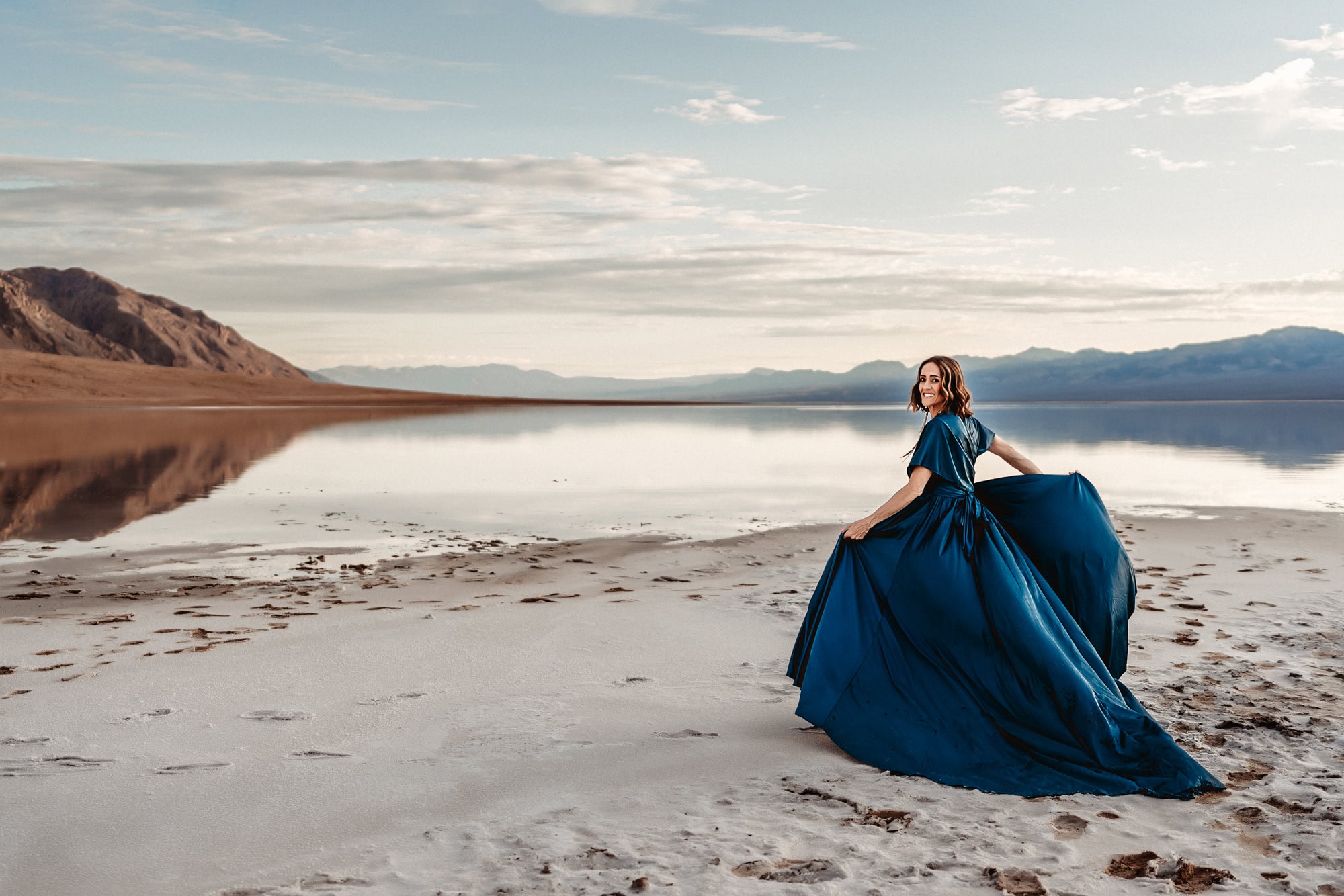 A woman wearing a dramatic blue dress twirls in it while looking at the camera during a branding photo shoot in Death Valley National Park's Badwater Basin. 