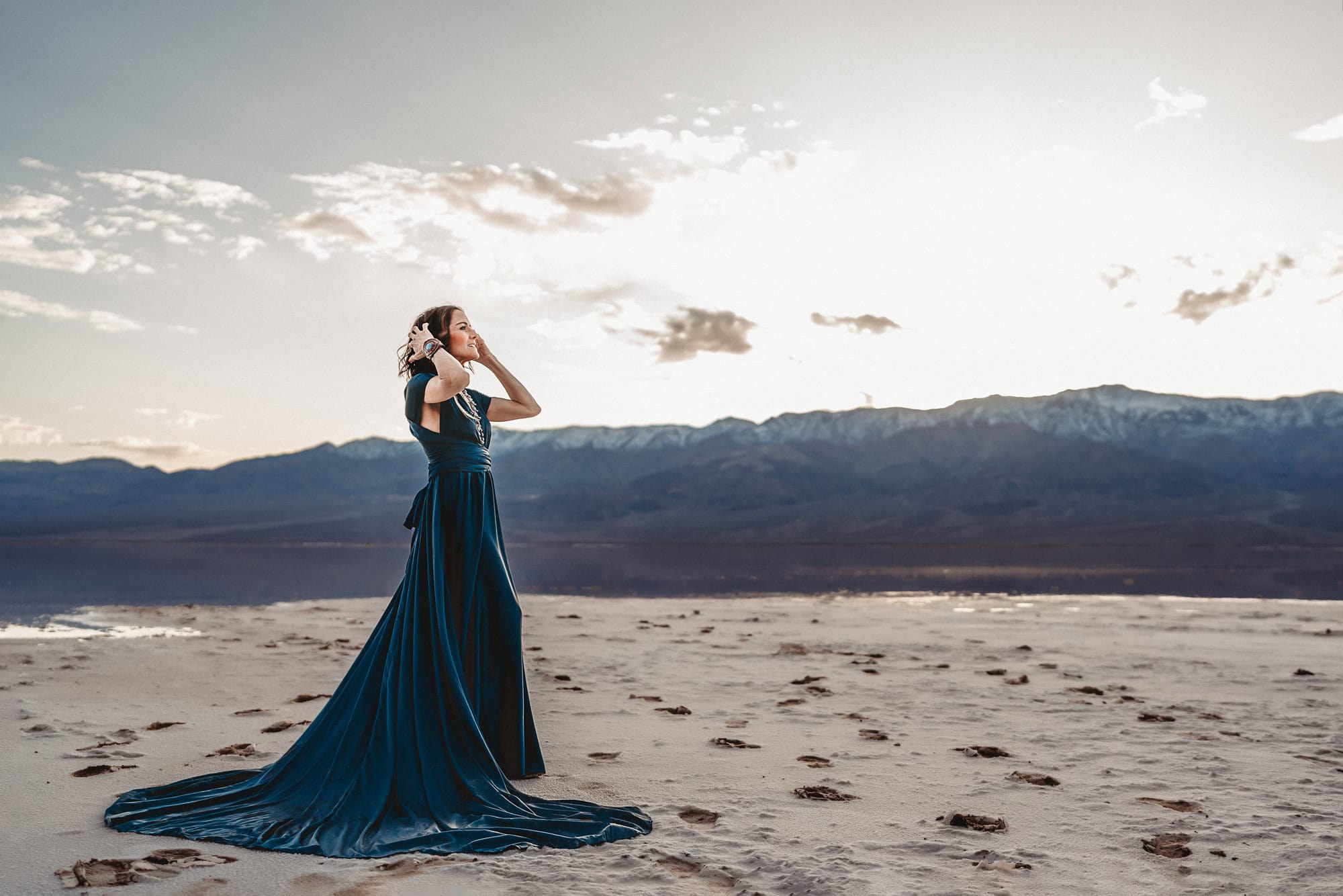 A woman wearing a dramatic blue dress with a long train stands in Death Valley National Park's Badwater Basin, touching her hair and looking off into the distance with the sun setting behind her. 