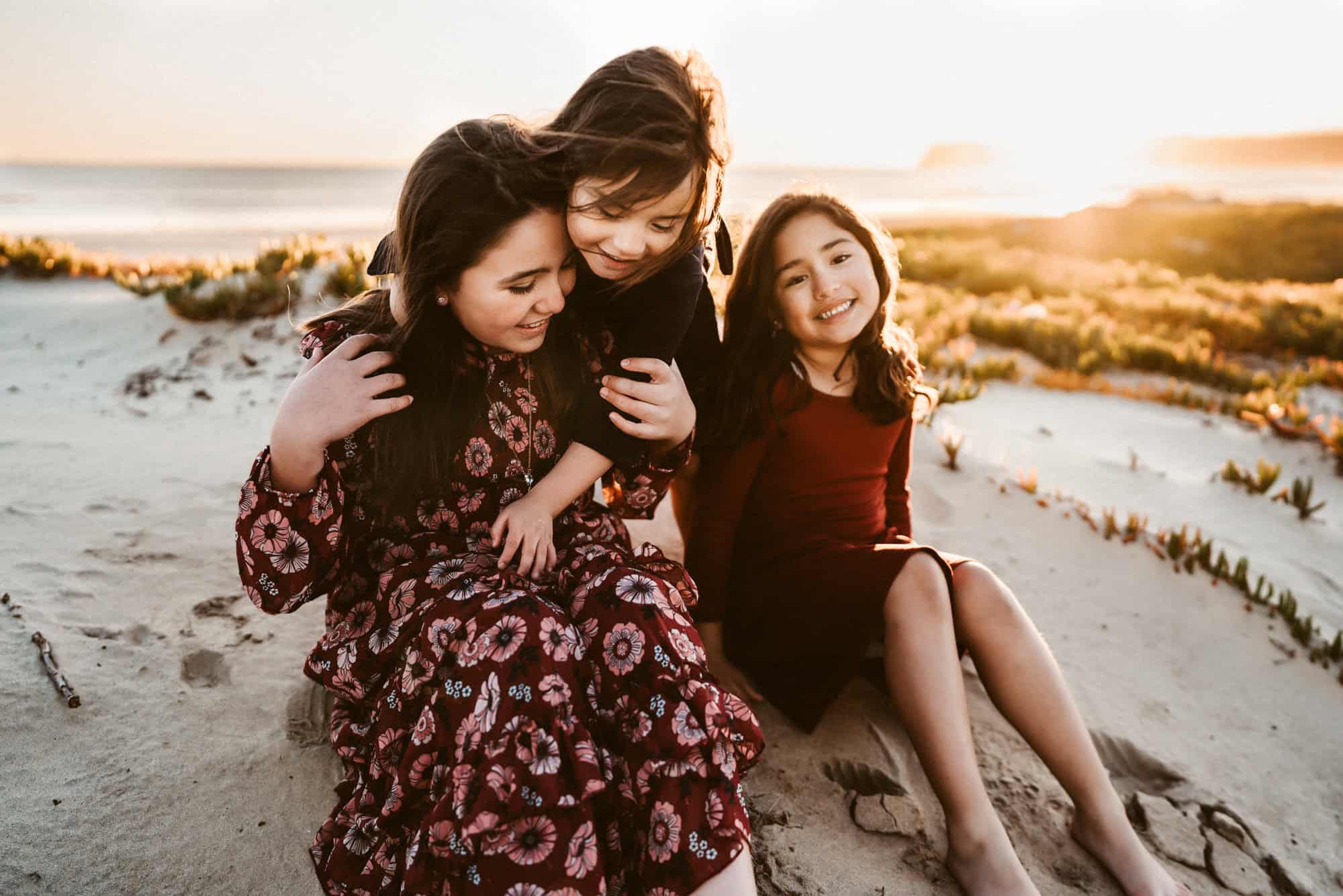 Sisters sitting together and laughing during a golden hour family photoshoot at Coronado Beach in San Diego by Love Michelle Photography.