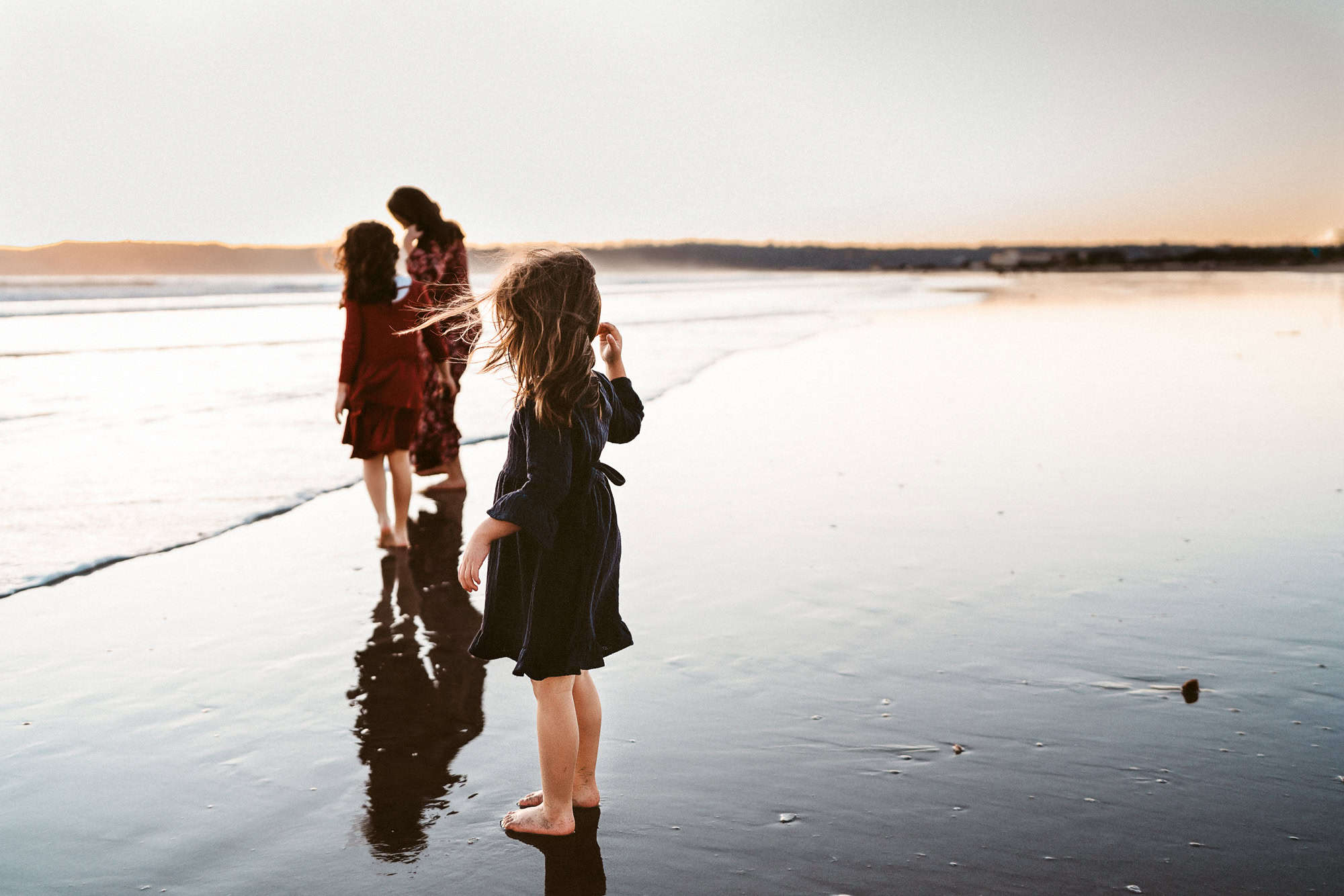 3 little girls stand on the shoreline of Coronado Beach at sunset, looking out to sea, during a San Diego beach photo session.