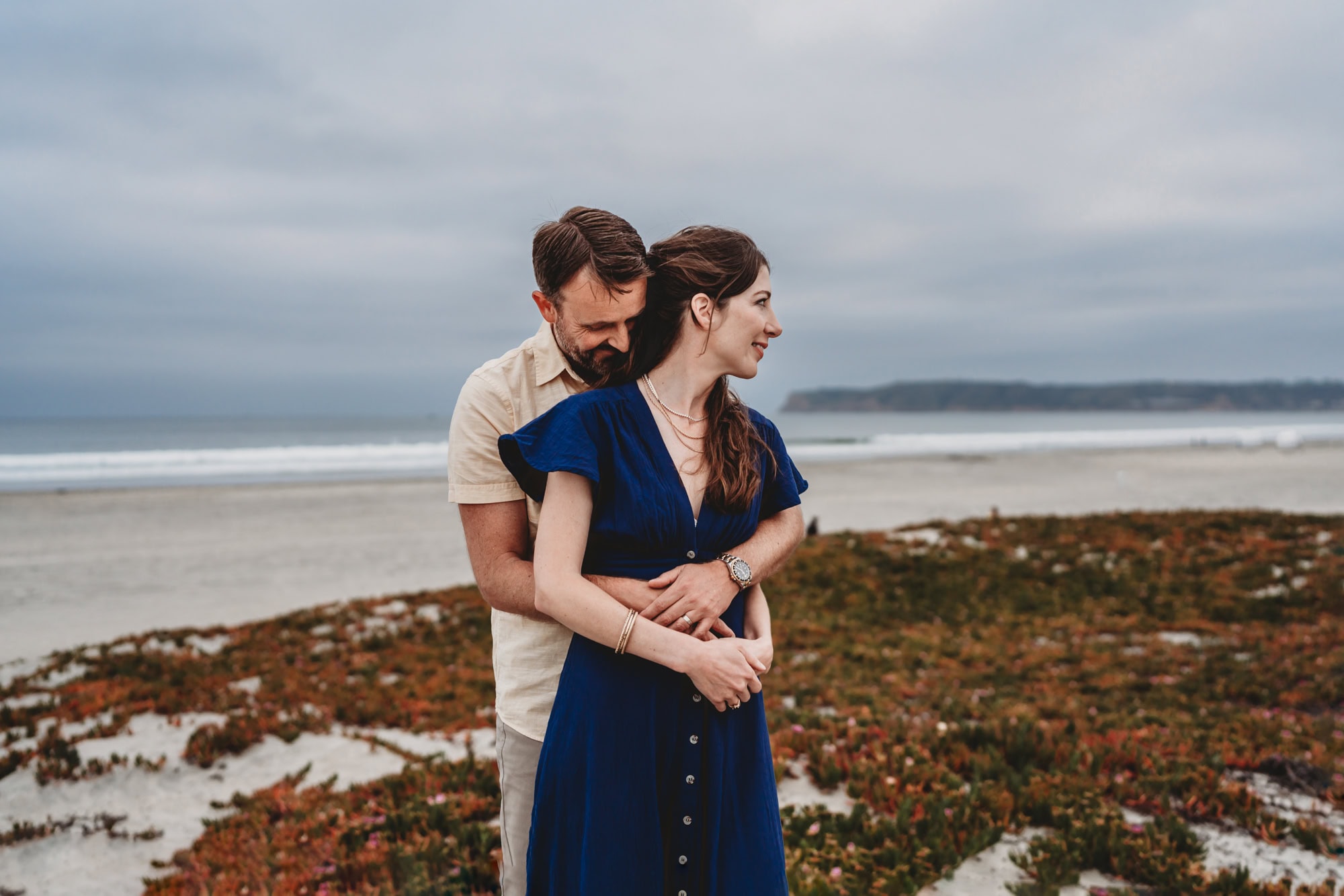 A man hugs his wife while she looks off to the side during a Coronado Beach family photoshoot in San Diego. 