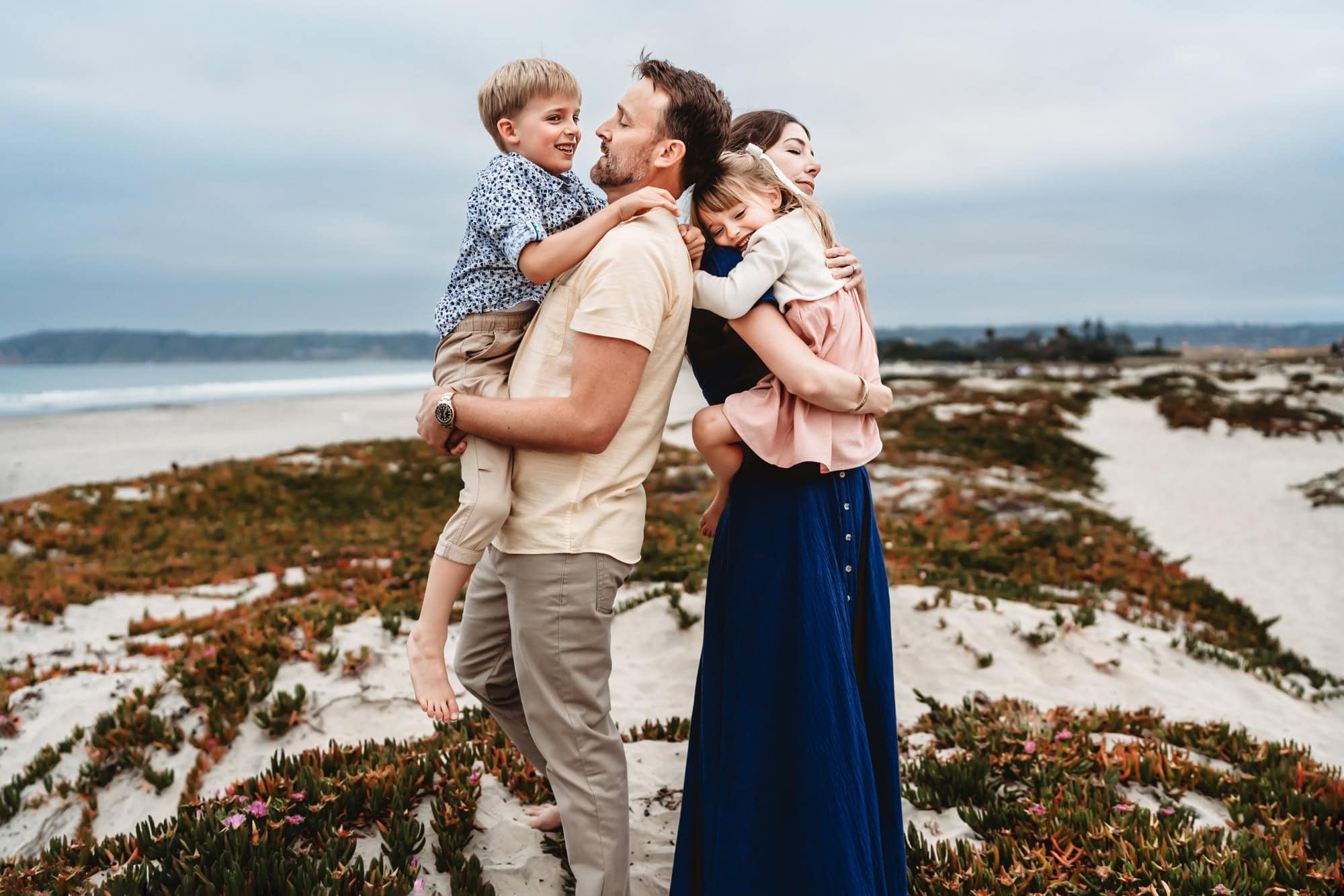 A man and woman stand back to back on a Coronado Beach sand dune, holding their children, during a San Diego family photoshoot.