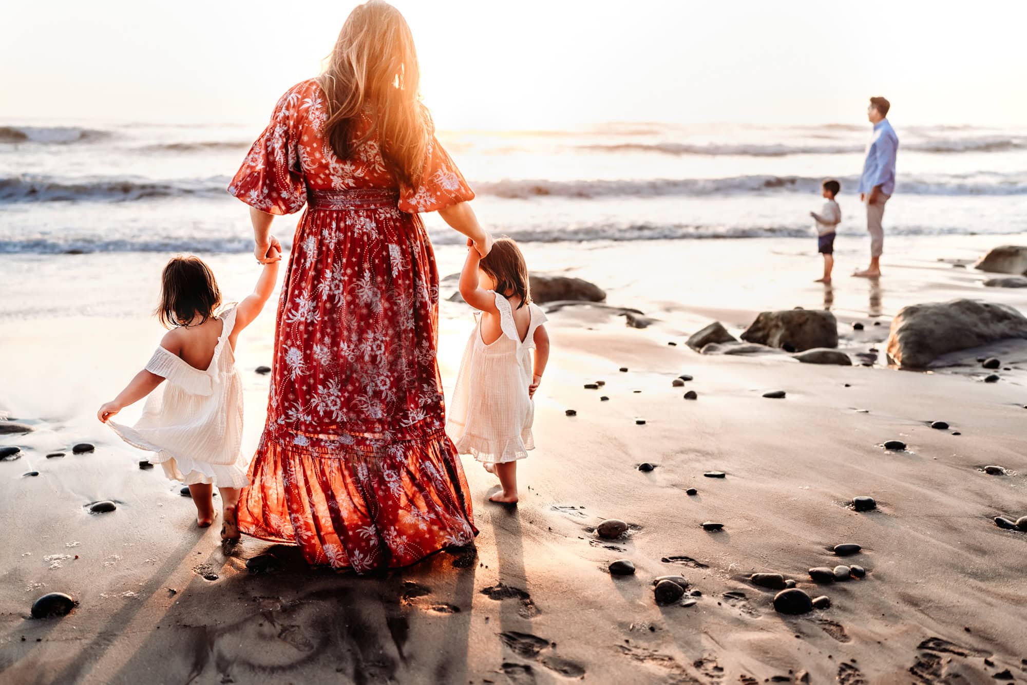 A woman wearing a long red dress holds her twin toddler daughters' hands while walking toward the shoreline of the beach in Del Mar, California. 