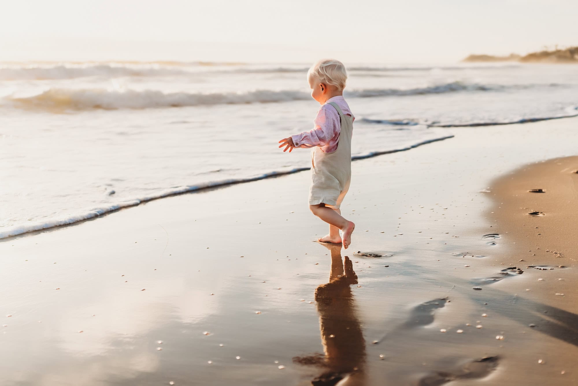 A blond toddler boy in white overalls runs toward the surf during a Carlsbad, California beach sunset.