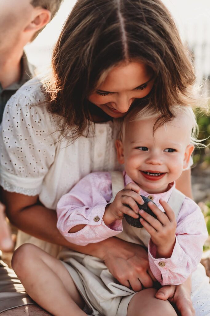 A mom sits on the ground, hugging her baby boy and smiling, during a family photoshoot in Carlsbad, CA. 