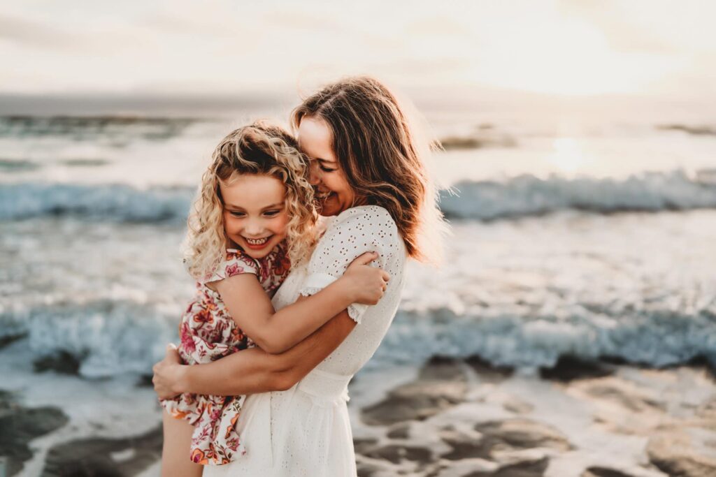 A mom wearing a white dress hugs her laughing 5 year old daughter on the beach in Carlsbad, CA.