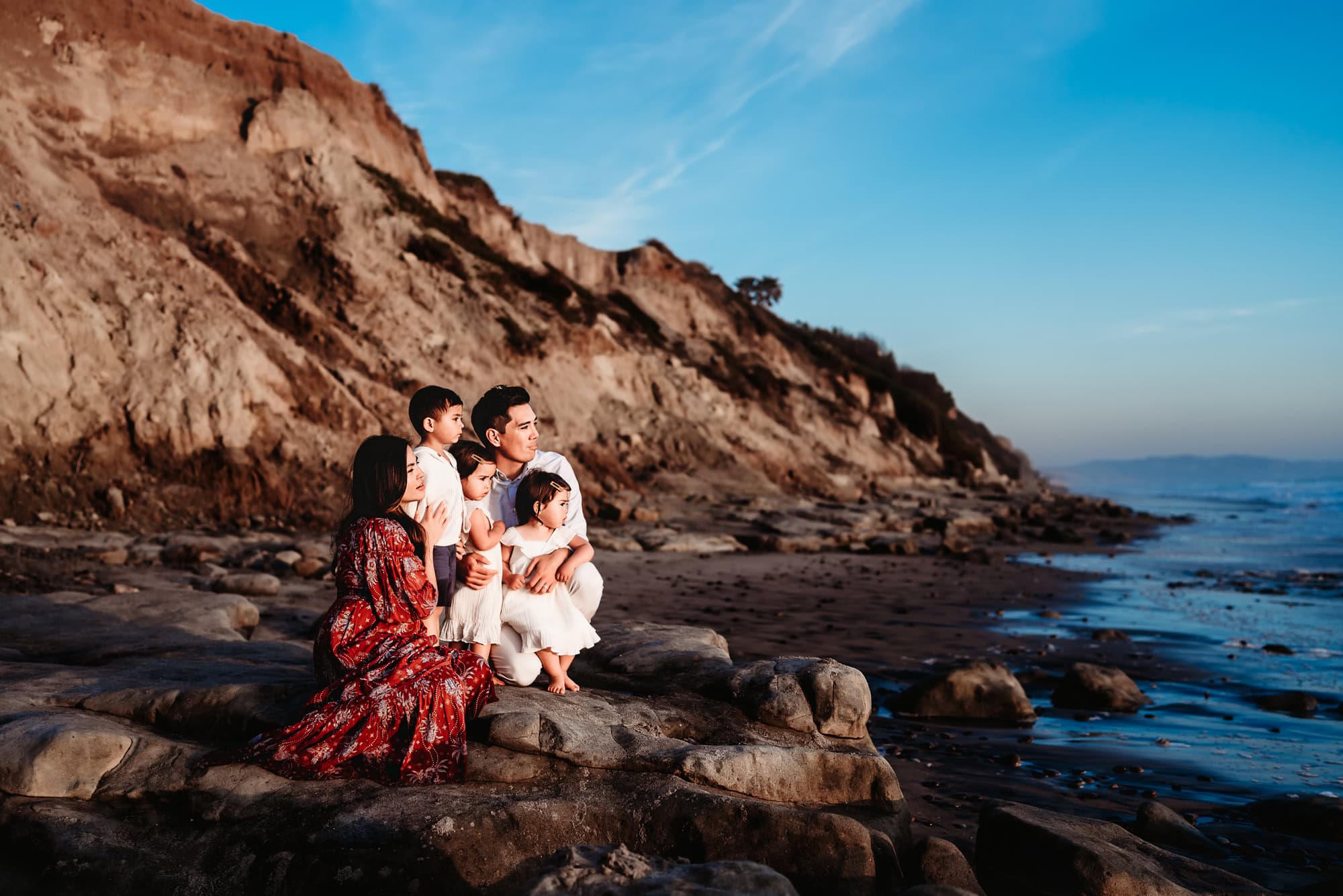 A family of 5 sits on the a rocky San Diego beach and enjoys the sunset during a lifestyle family photoshoot by Love Michelle Photography. 