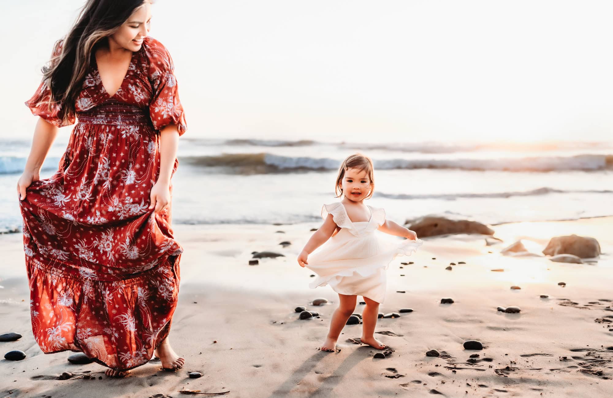 A toddler girl in a white dress holds her skirt and twirls around with her mama next to her during a lifestyle family photoshoot in Del Mar, CA