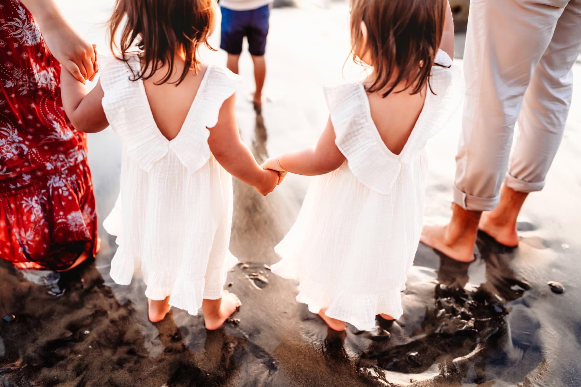 Twin toddler girls wearing matching white dresses hold hands during a family photoshoot on a Del Mar beach. 