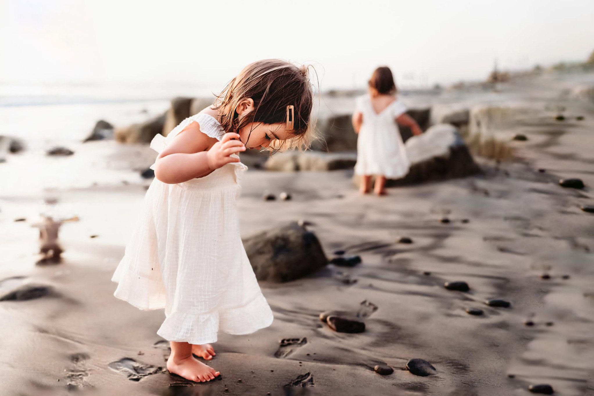 A toddler girl in a white dress stands on the beach in Del Mar, California, looking at the sand around her feet.