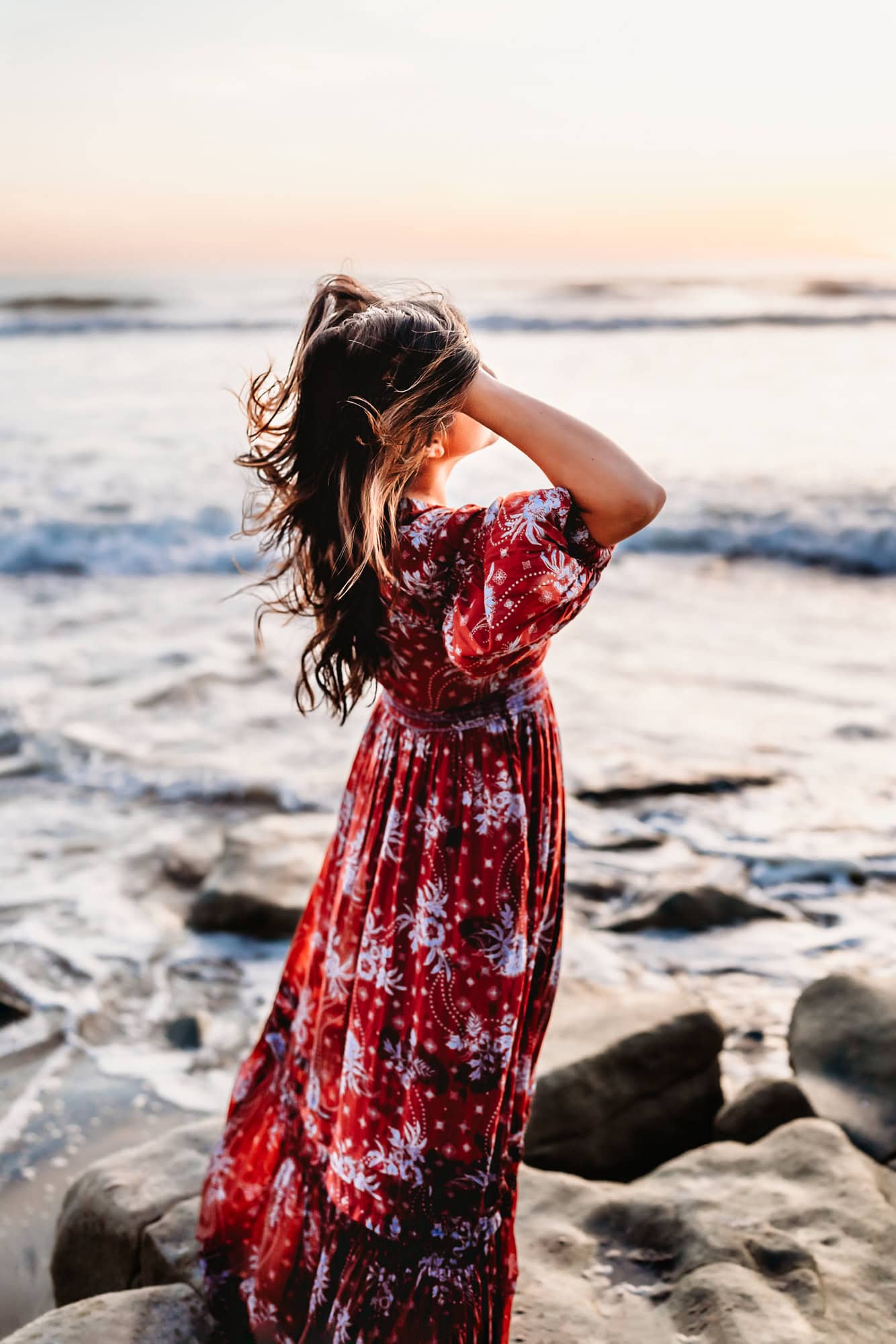 A woman in a red Free People maxi dress tosses her hair back while watching the sunset on a rocky San Diego beach. 