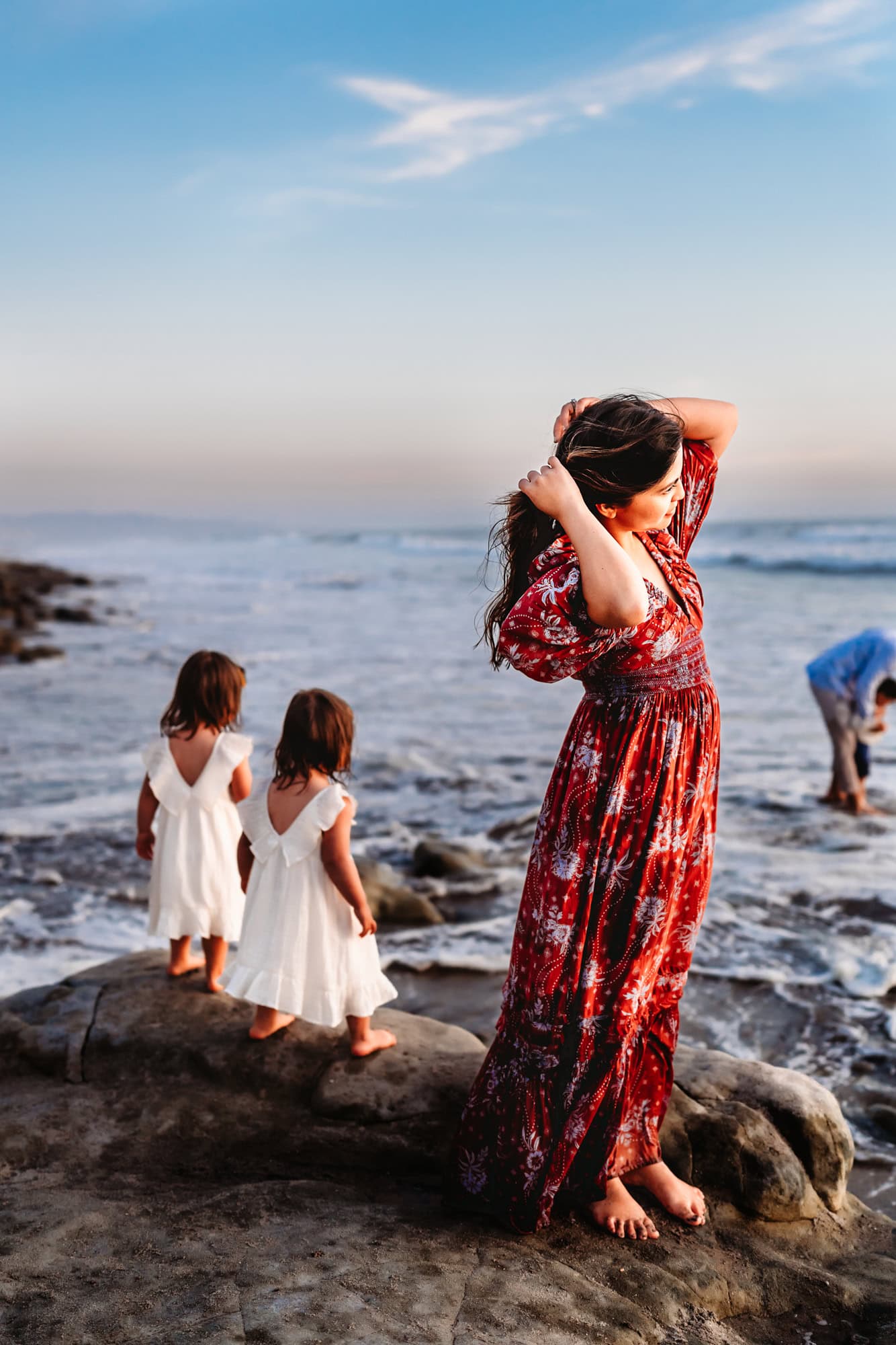 A woman in a red Free People maxi dress plays with her hair  while watching the sunset on a rocky Del Mar beach. Her twin toddler girls stand behind her, looking at the water. 