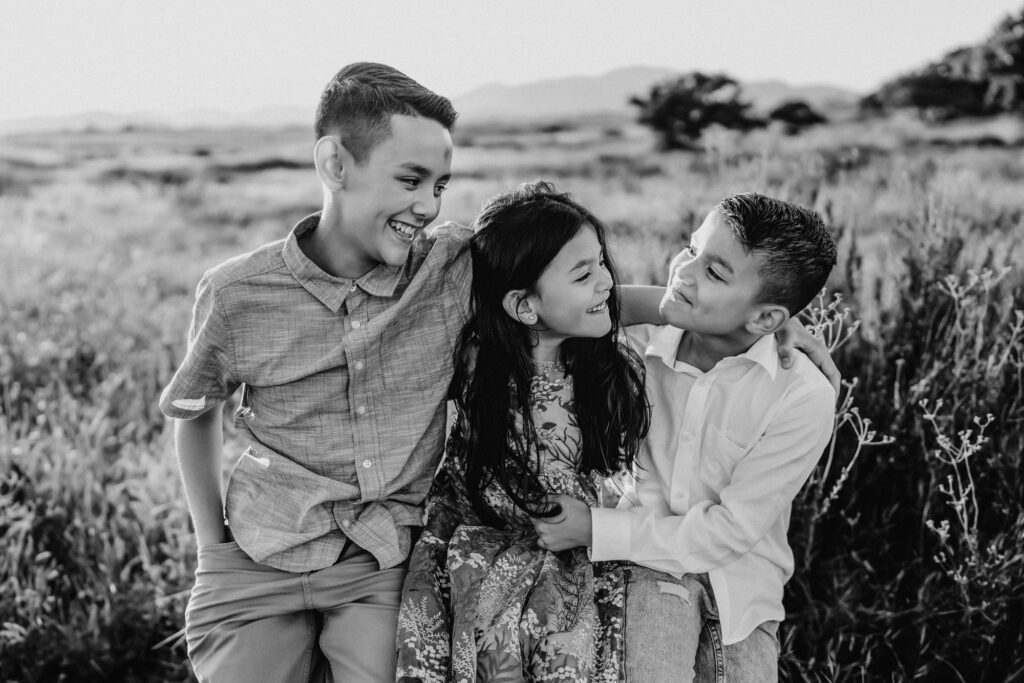 Three young siblings sit on a large rock, looking at and laughing with each other during a lifestyle family photo session in Alpine, California. 