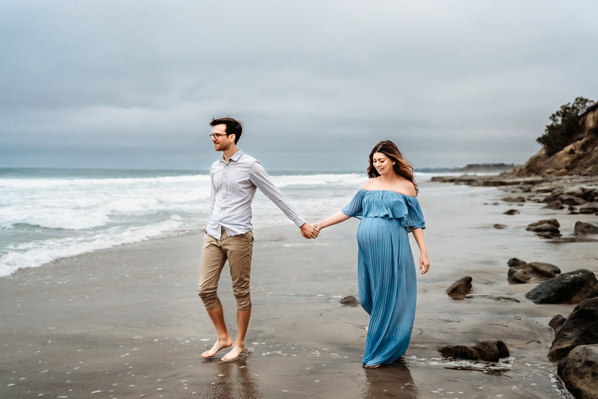 A man and his pregnant wife hold hands and walk on the beach in Del Mar, CA, during a lifestyle maternity photo session. 