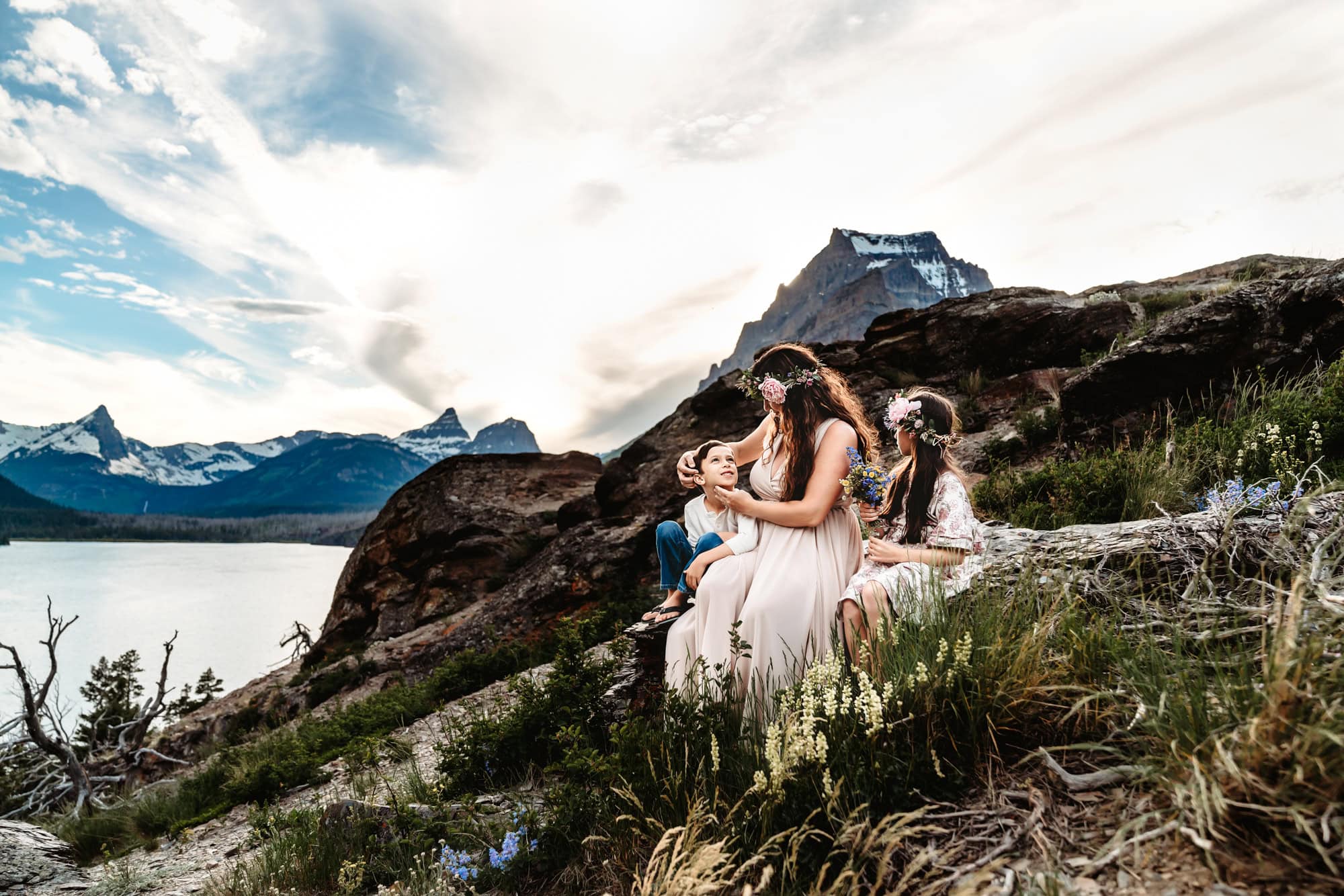 A mom sits with her young son and daughter above Montana's St. Mary's Lake during a Glacier National Park lifestyle family photo session by Love Michelle Photography.