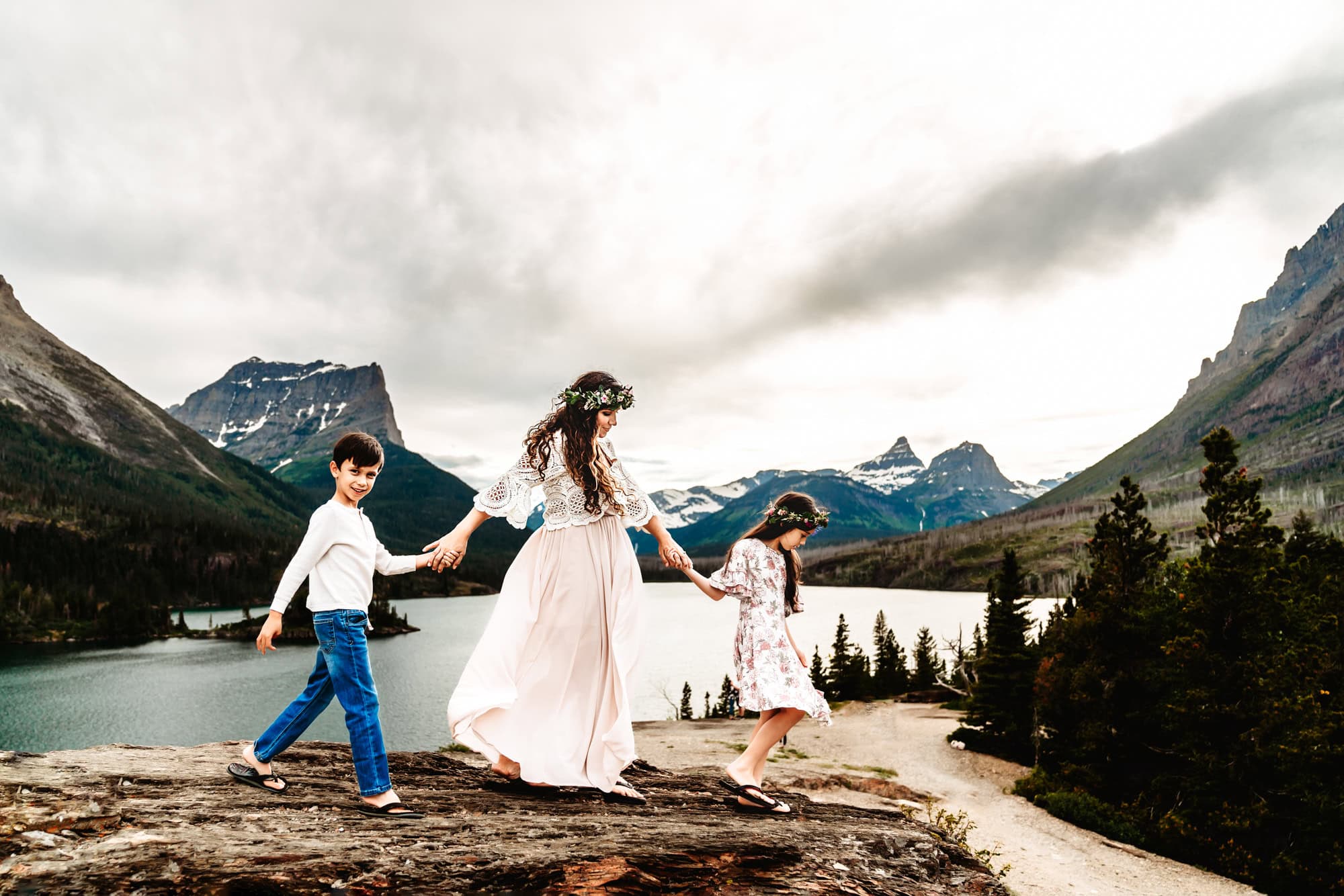 A mom holds hand with her two kids and walks on a large rock during a family photo shoot in Glacier National Park.