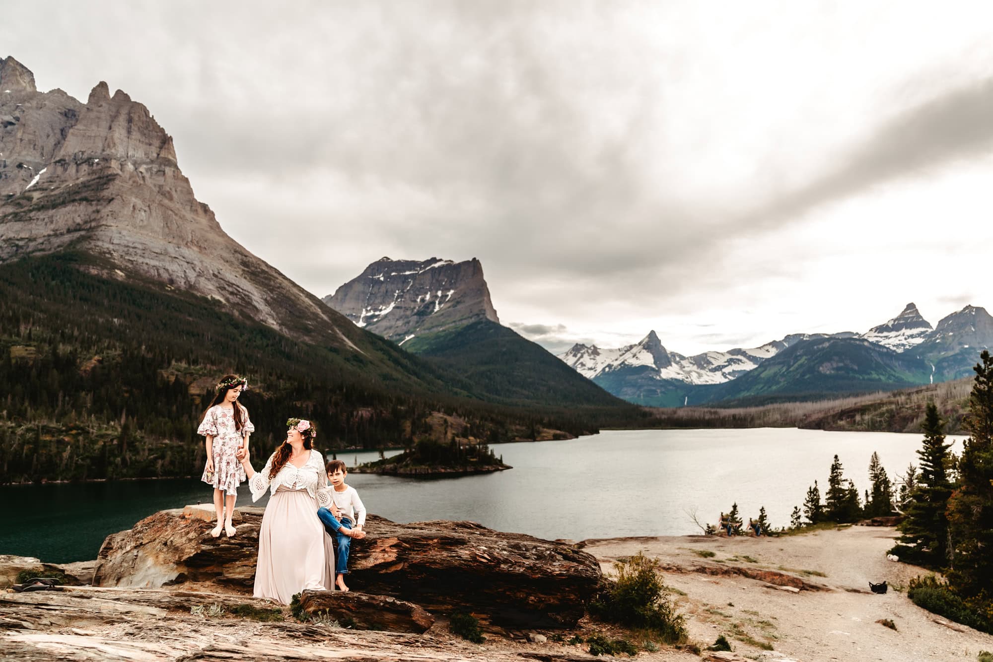 A mom sits on a large boulder in Glacier National Park, looking at her children and smiling. St. Mary's Lake lies in the background at the base of multiple mountain peaks. 