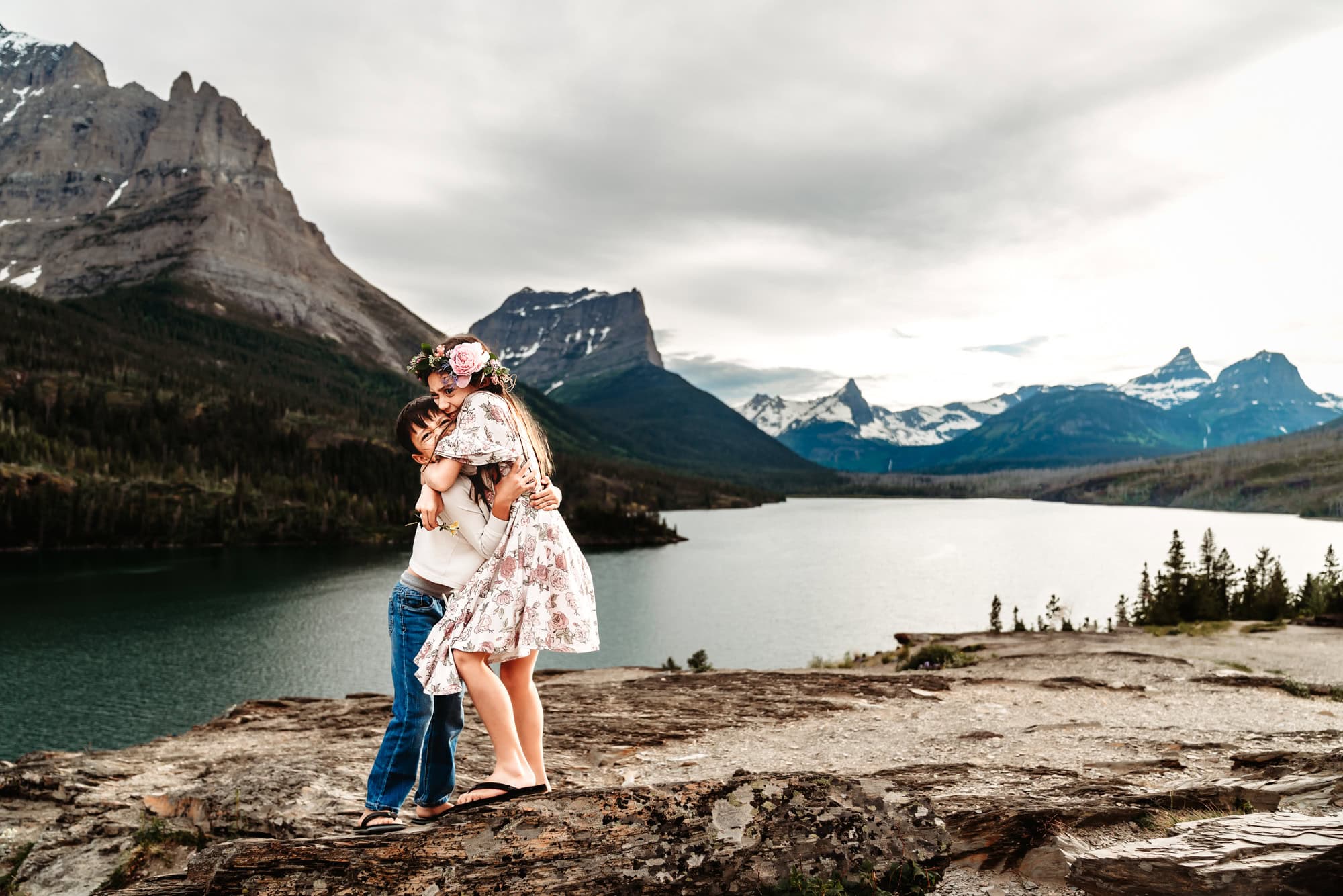 A young boy and his sister embrace on a rocky ledge above Glacier National Park's St. Mary's Lake during a family photo session. 