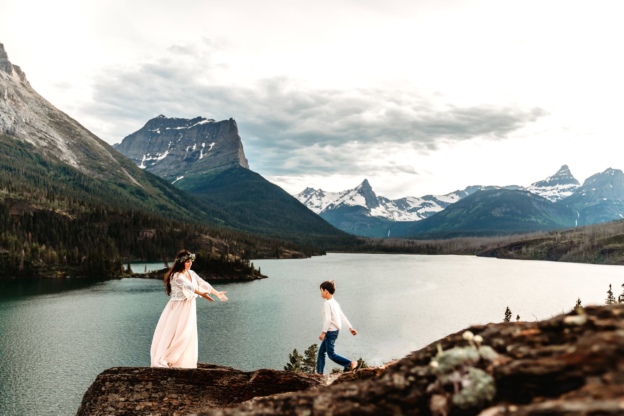 A mother in a long dress stands on a rock ledge above St. Mary's Lake and reaches out to her young son who is running to meet her. 