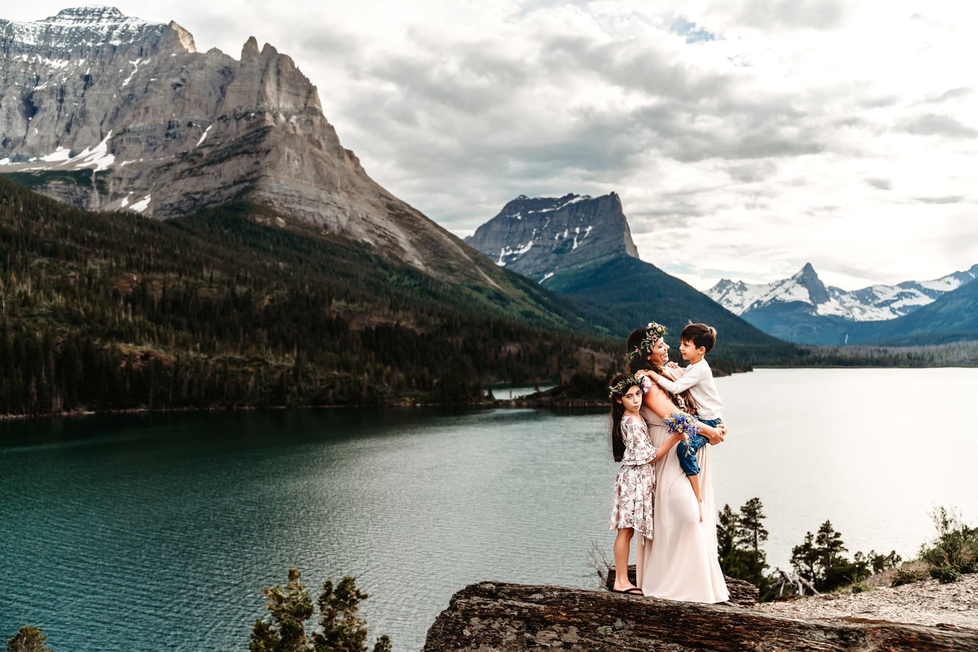 A mom in a long dress embraces her two children while standing on a rocky ledge above Glacier National Park's St. Mary's Lake. 