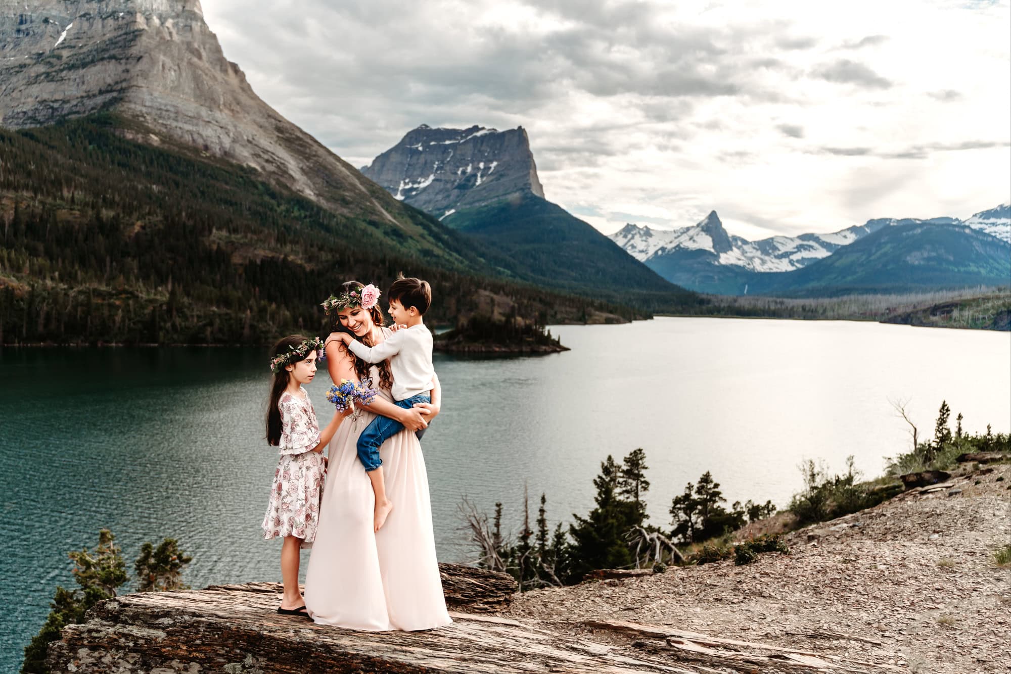 A mom in a long dress embraces her two children while standing on a rocky ledge during a lifestyle family photoshoot. She's standing above Glacier National Park's St. Mary's Lake in St. Mary, Montana. 