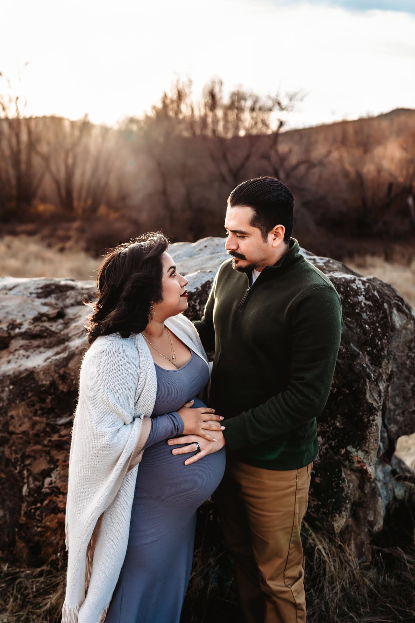 A couple in San Diego stand facing one another in front of a large boulder in the Cuyamaca Mountains. She is pregnant and he has placed his hand on her belly. 