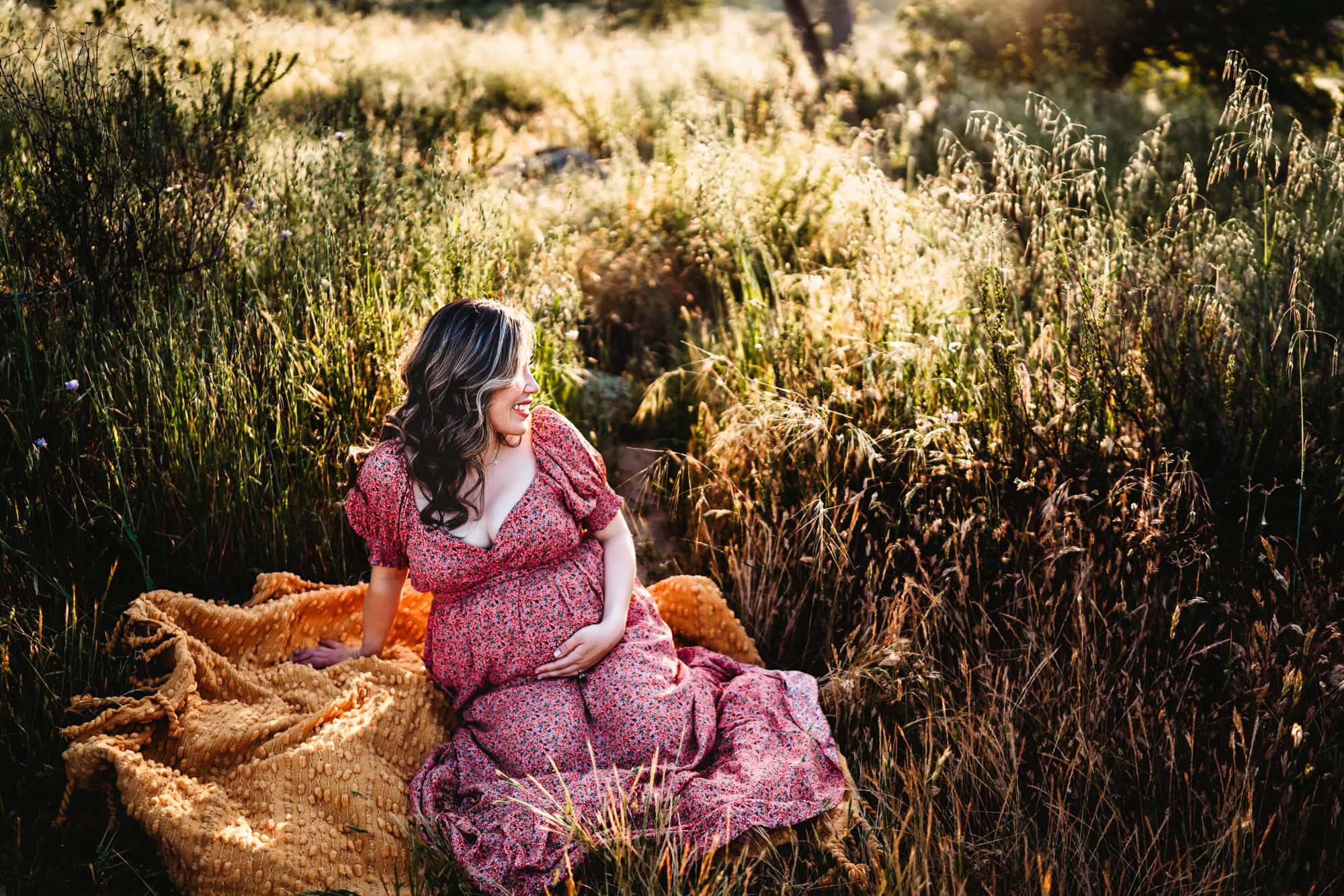 A pregnant woman in a long pink dress hold her belly, smiles, and looks off to her left while sitting on a blanket in a San Diego field. 