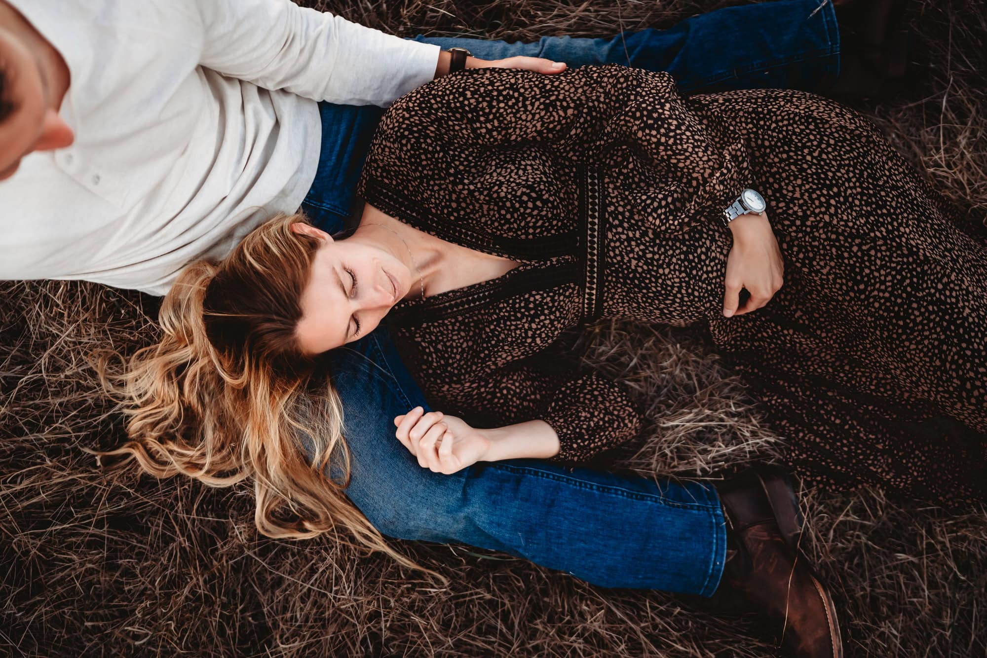 A blonde pregnant woman wearing a long brown dress lays her head on her husband's lap. Both are in a rustic field during a San Diego lifestyle maternity photoshoot in Mission Trails Regional Park 