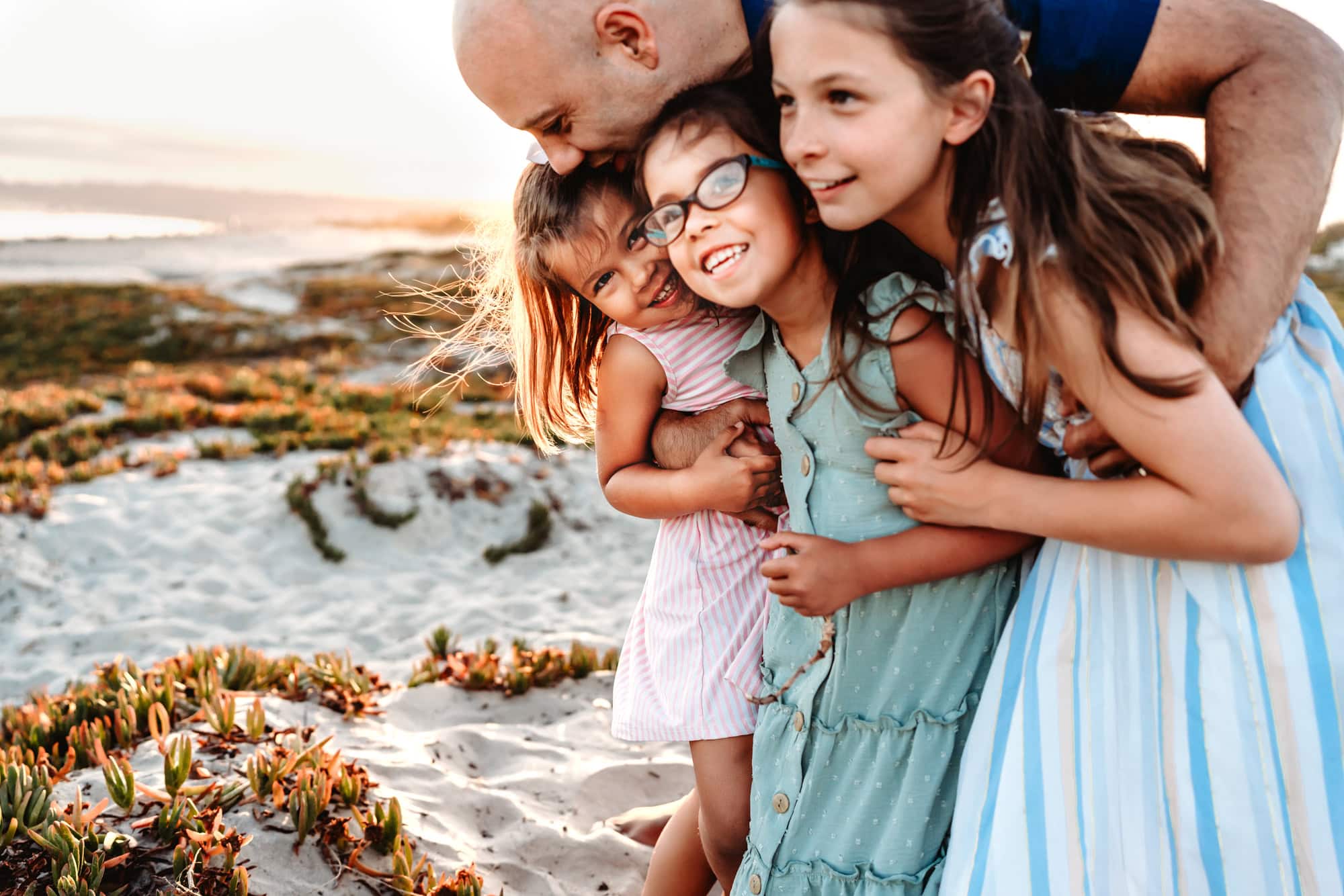A fasther standing on Coronado Beach, leaning over and hugging his three young daughters, who are all looking at toward the ocean and laughing.
