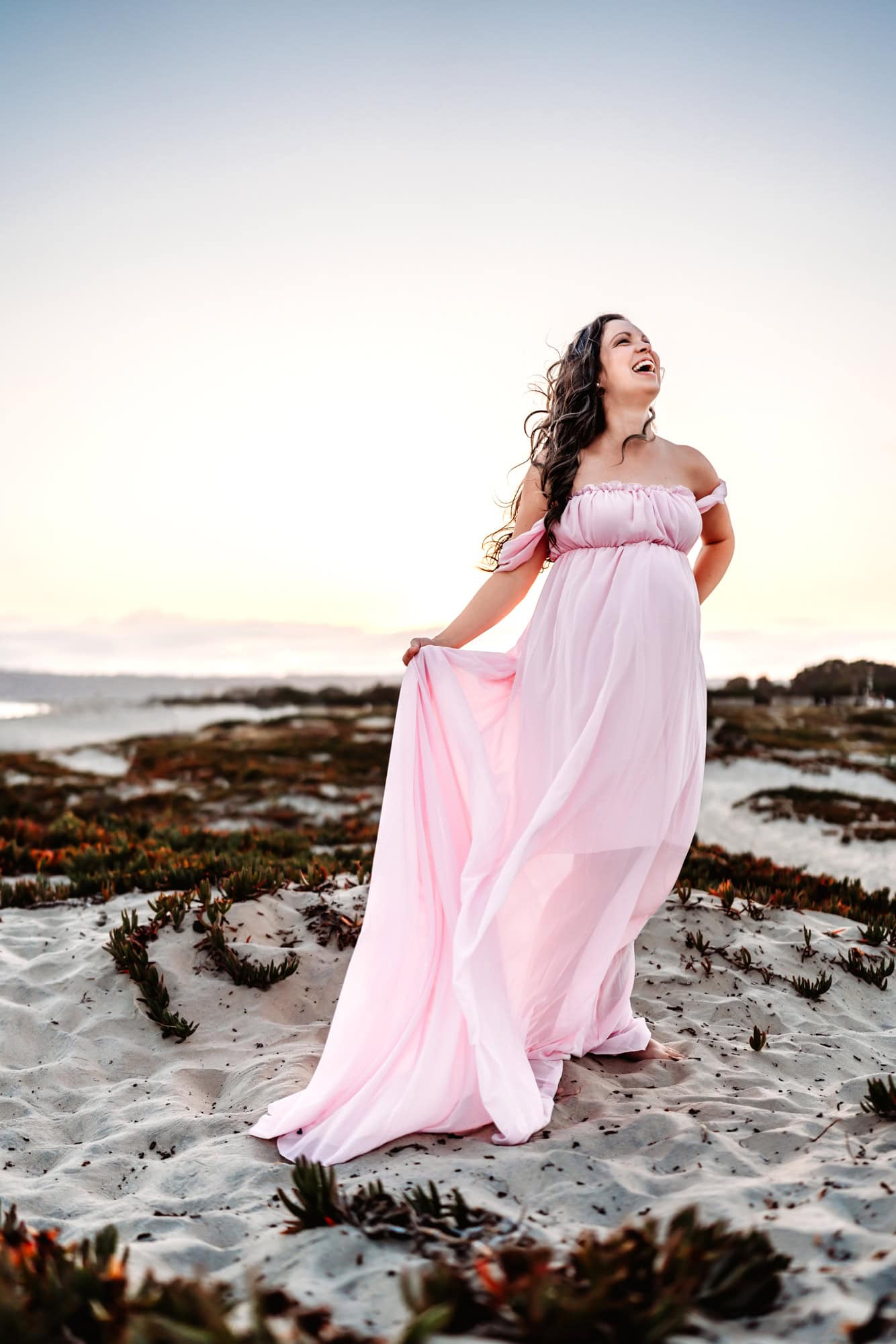 A brunette pregnant woman stands on a beach, holding the skirt of her long and flowy pink dress and laughing toward the sky. 