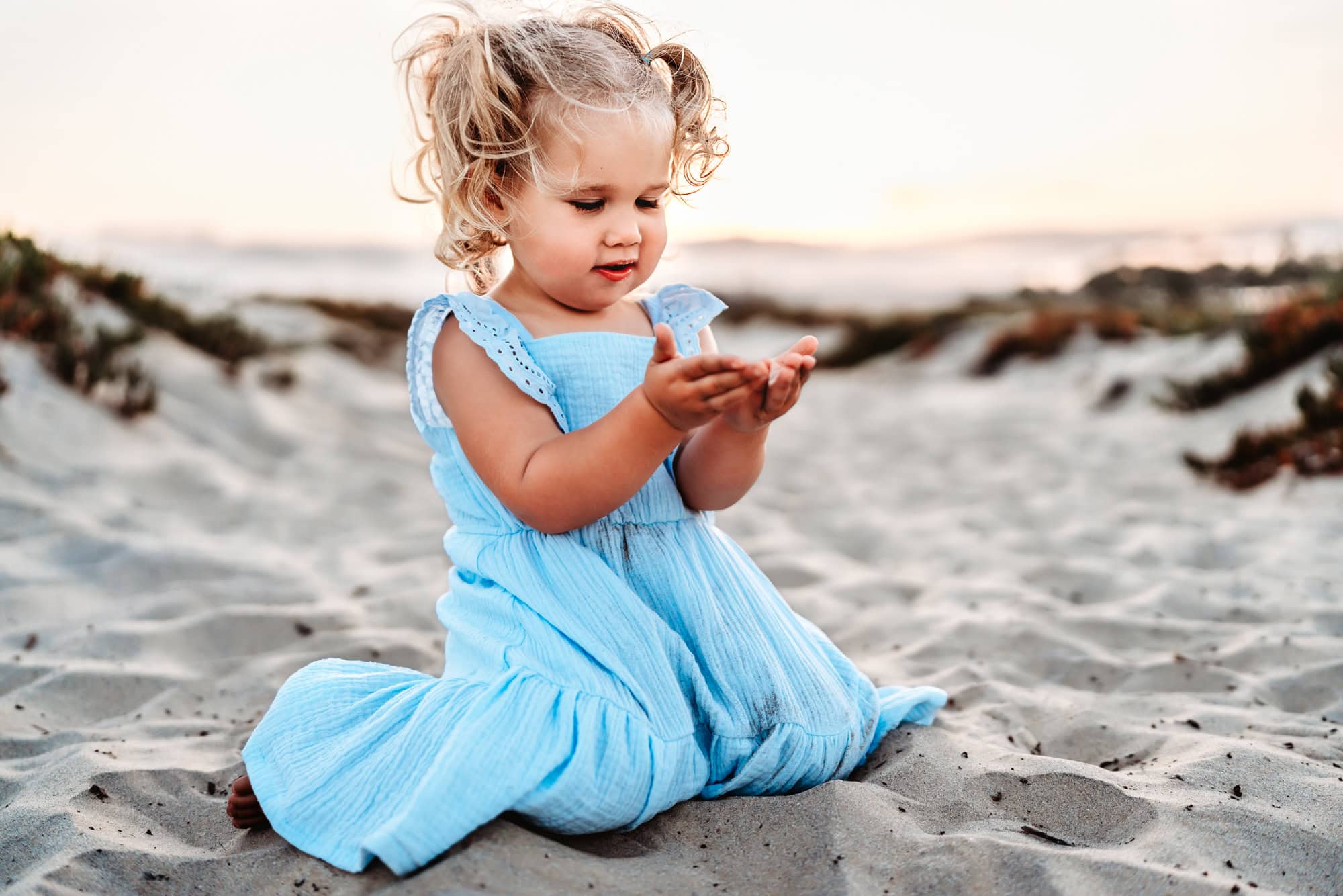 A little blond toddler girl wearing a blue dress sits in the sand and looks at the sand she's holding in her hands. 