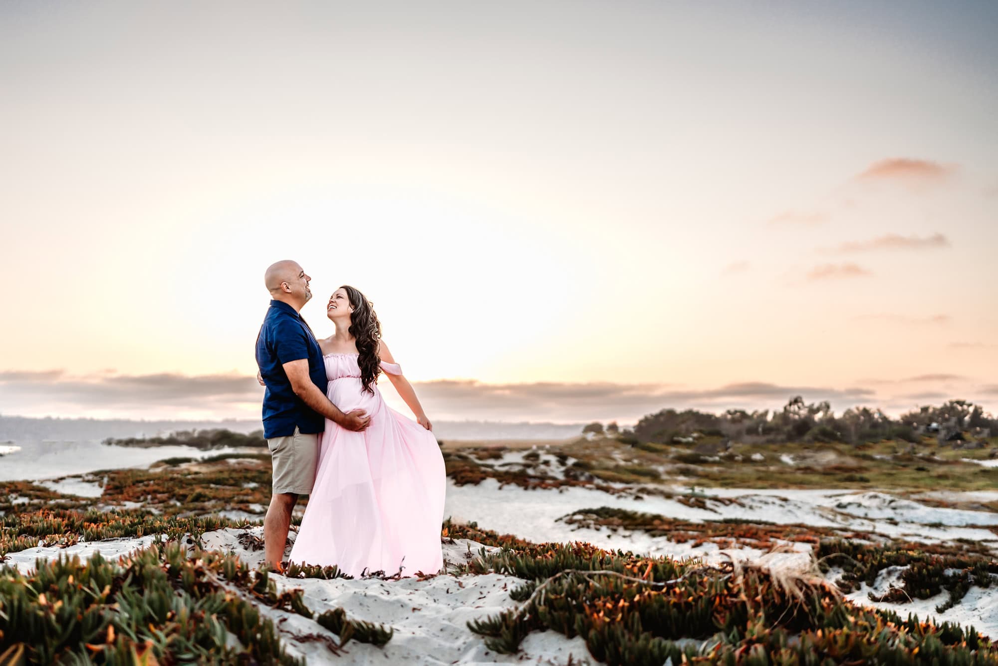 A pregnant woman wearing a flowy pink dress stands on a San Diego beach sand dune at sunset and embraces her husband, in an article listing reasons to book a San Diego lifestyle maternity photoshoot. 