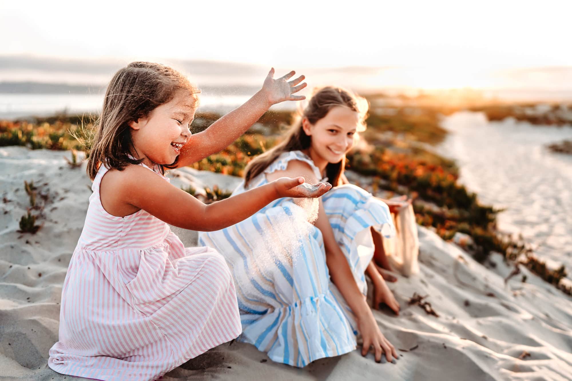 A little girl sits with her sister on a Coronado Island sand dune and laughs as sand sifts through her fingers, backlit by the sun. 