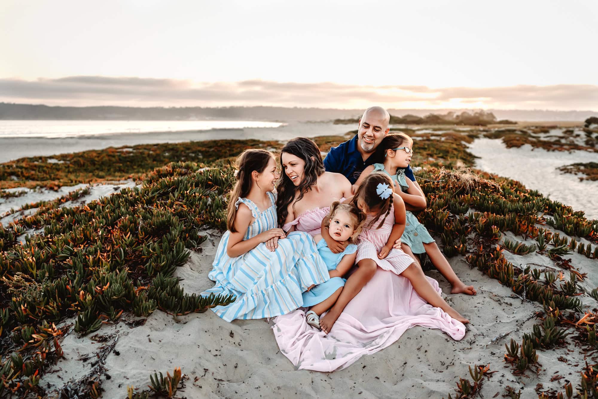 A family of 6 sits on a Coronado Island sand dune during a beach family photoshoot, smiling and looking at each other. 