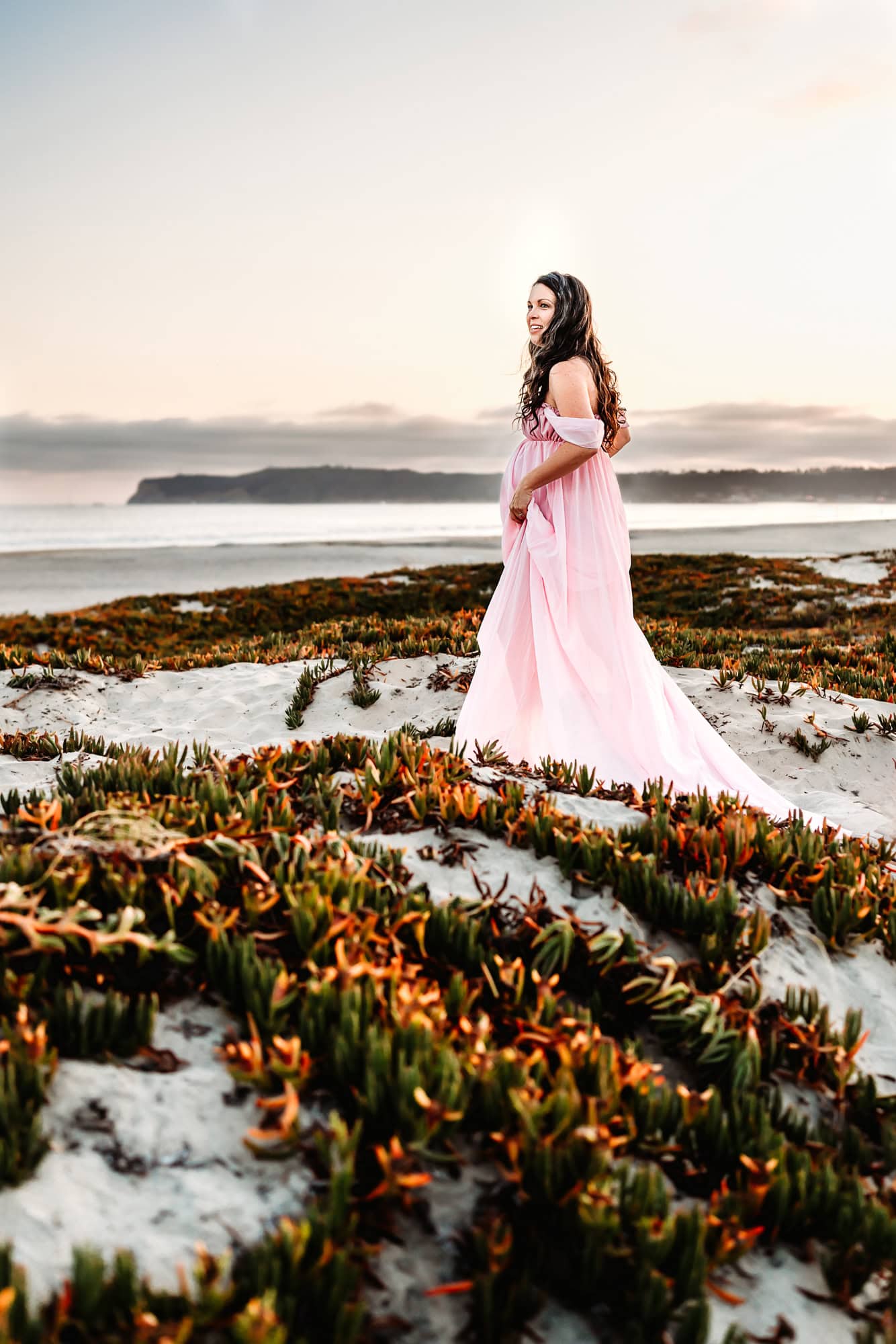 A pregnant woman with long dark hair, wearing a flowy pink dress, holds her skirts up and walks on a San Diego sand dune during a maternity photoshoot. 