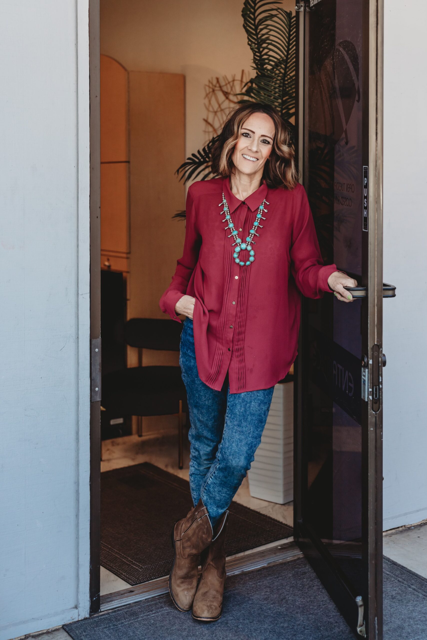 A woman wearing a red shirt, jeans, and boots stands in an open doorway, smiling. 