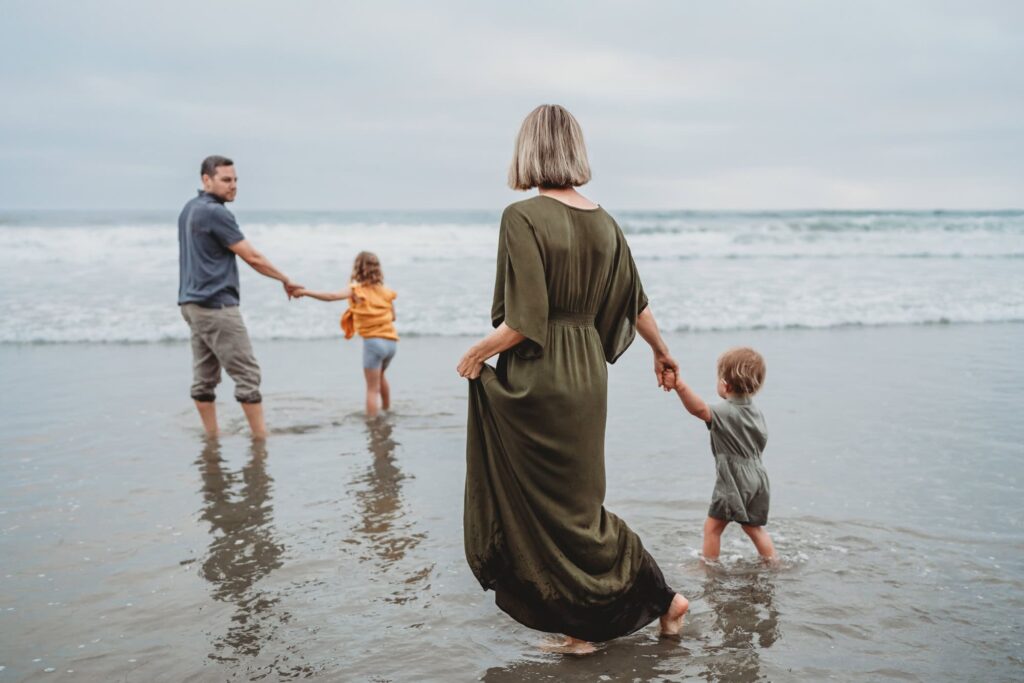 San Diego Family Photographer captures family standing on beach holding hands while tide rolls in and out in San Diego