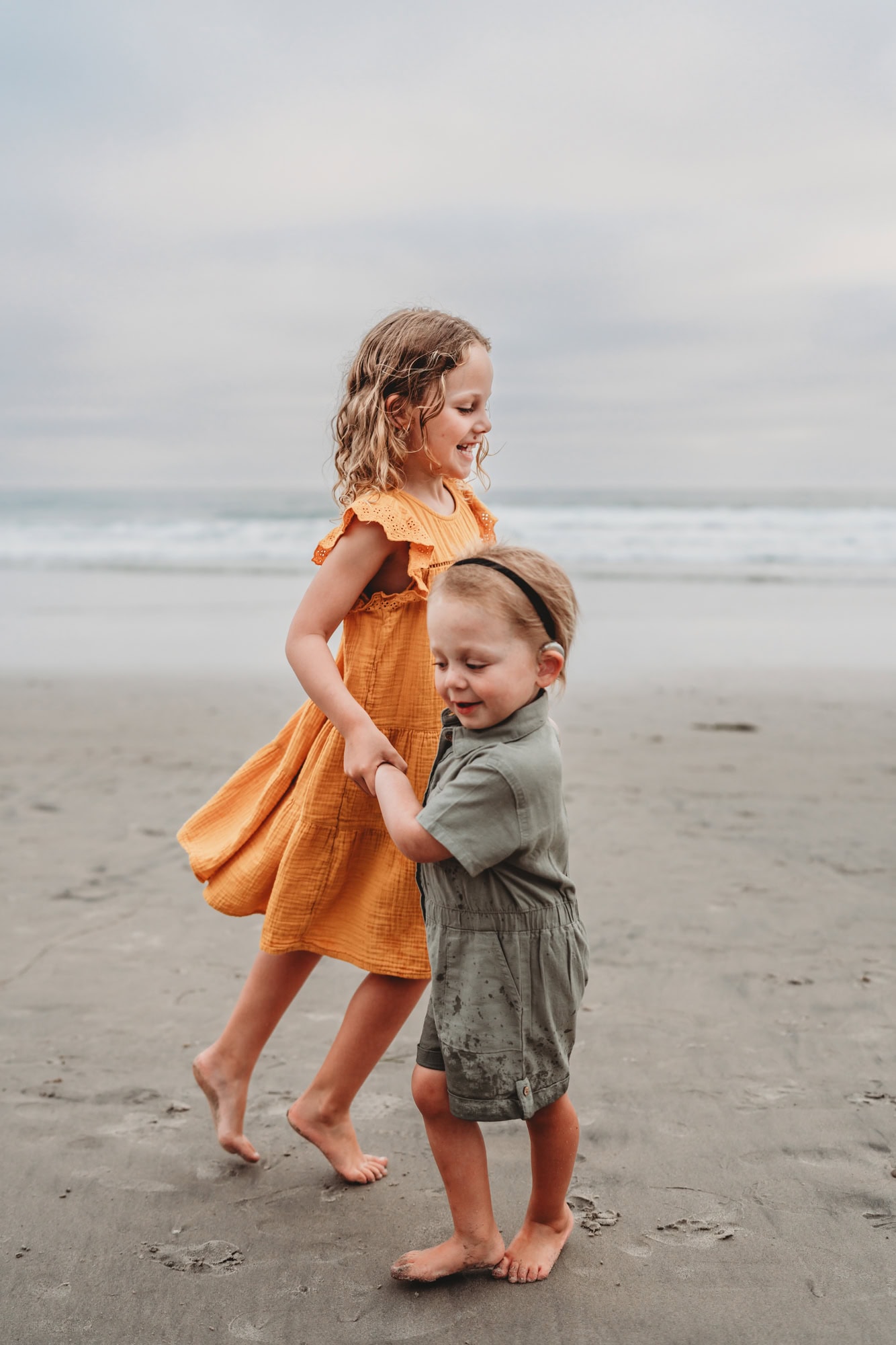 A girl in a yellow dress holds hands with her little sister while playing "ring around the Rosie" on the beach in Del Mar, CA. 