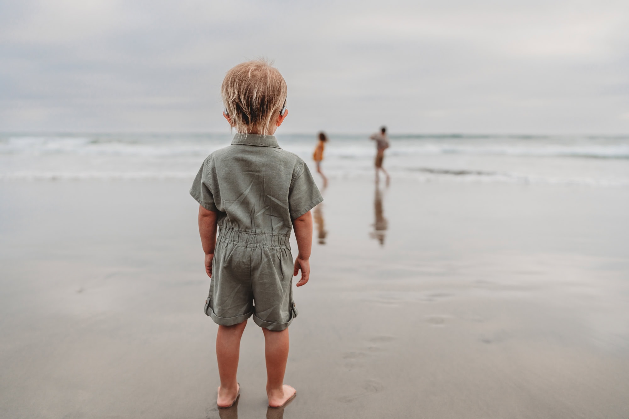 A little girl stands on a Del Mar beach, looking at her siblings playing in the surf. 