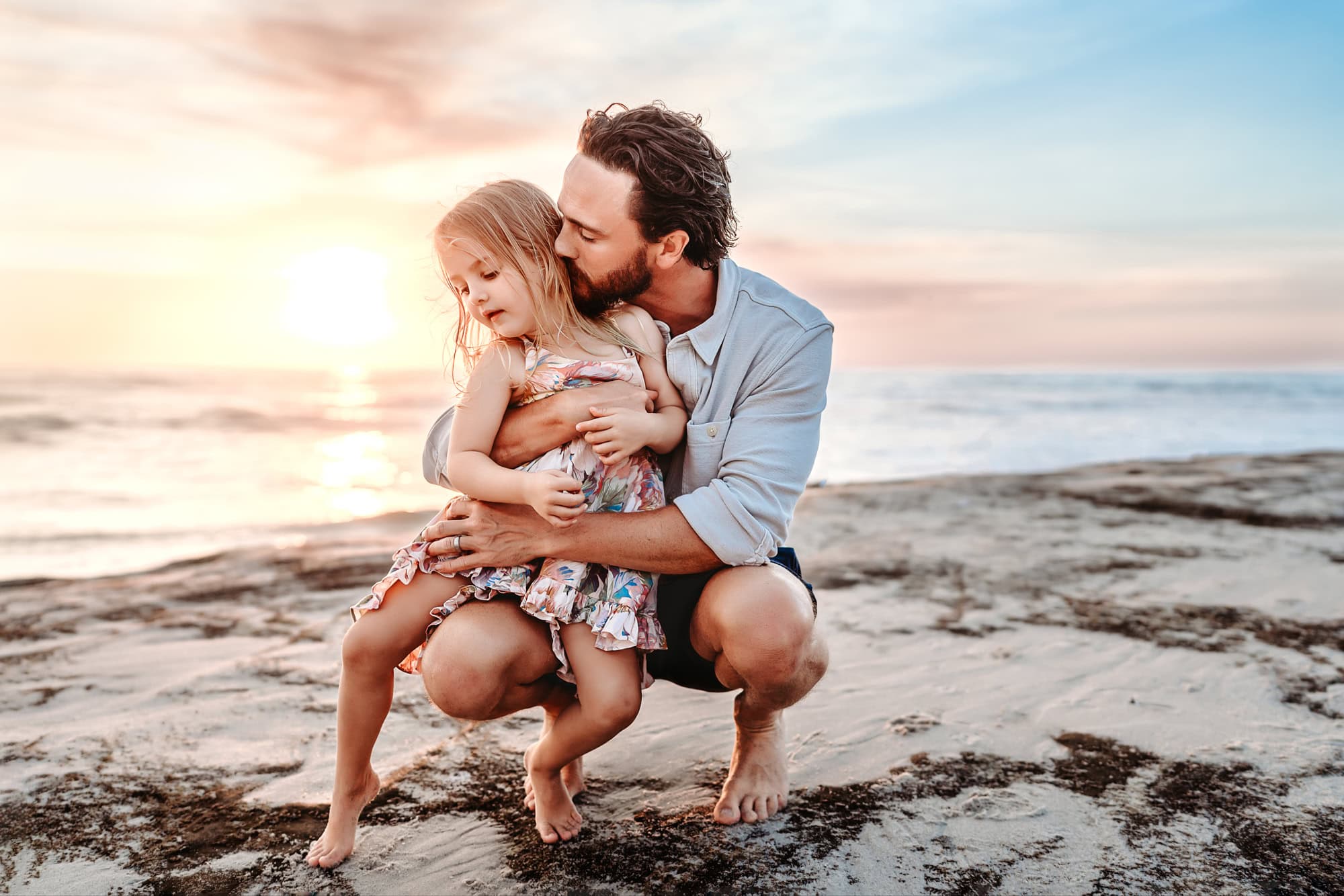 A man crouches on a rock, snuggling his little girl, during a La Jolla beach family photo shoot. 