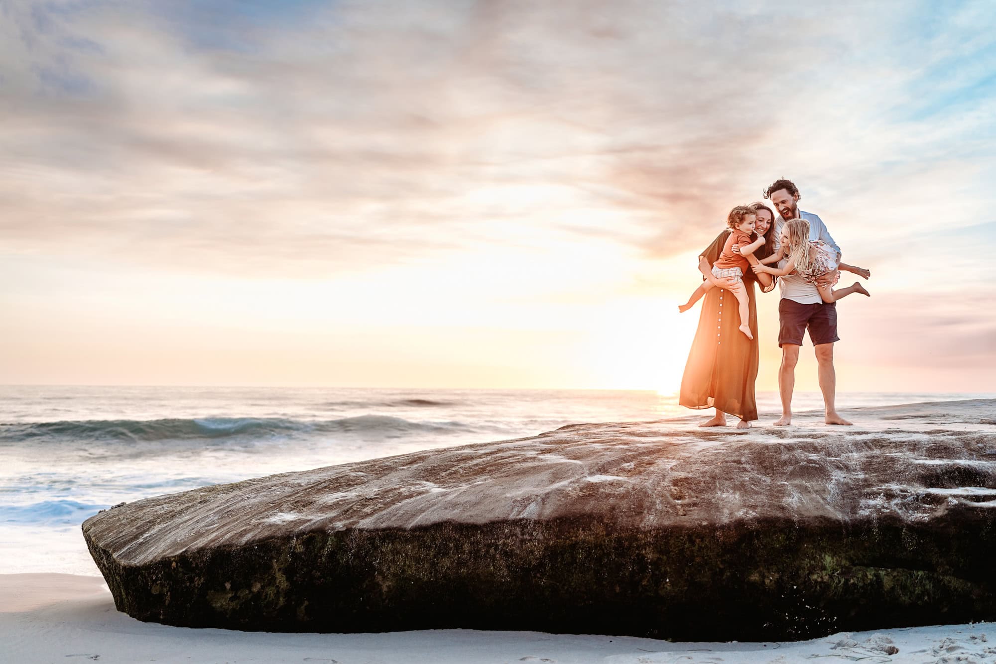 A family of four stands on a large rock during a sunset family photoshoot on La Jolla's Windansea Beach in California.