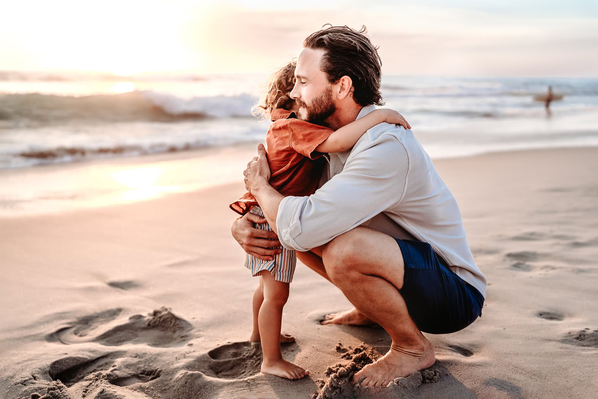 A man crouches down in the sand, hugging his two year old son, during a sunset beach photo shoot in La Jolla, California.