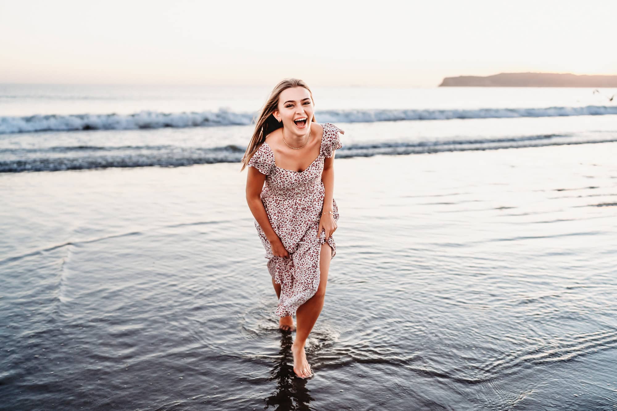 A young woman holds her skirt, laughing, and run out of the surf on Coronado Beach in San Diego.